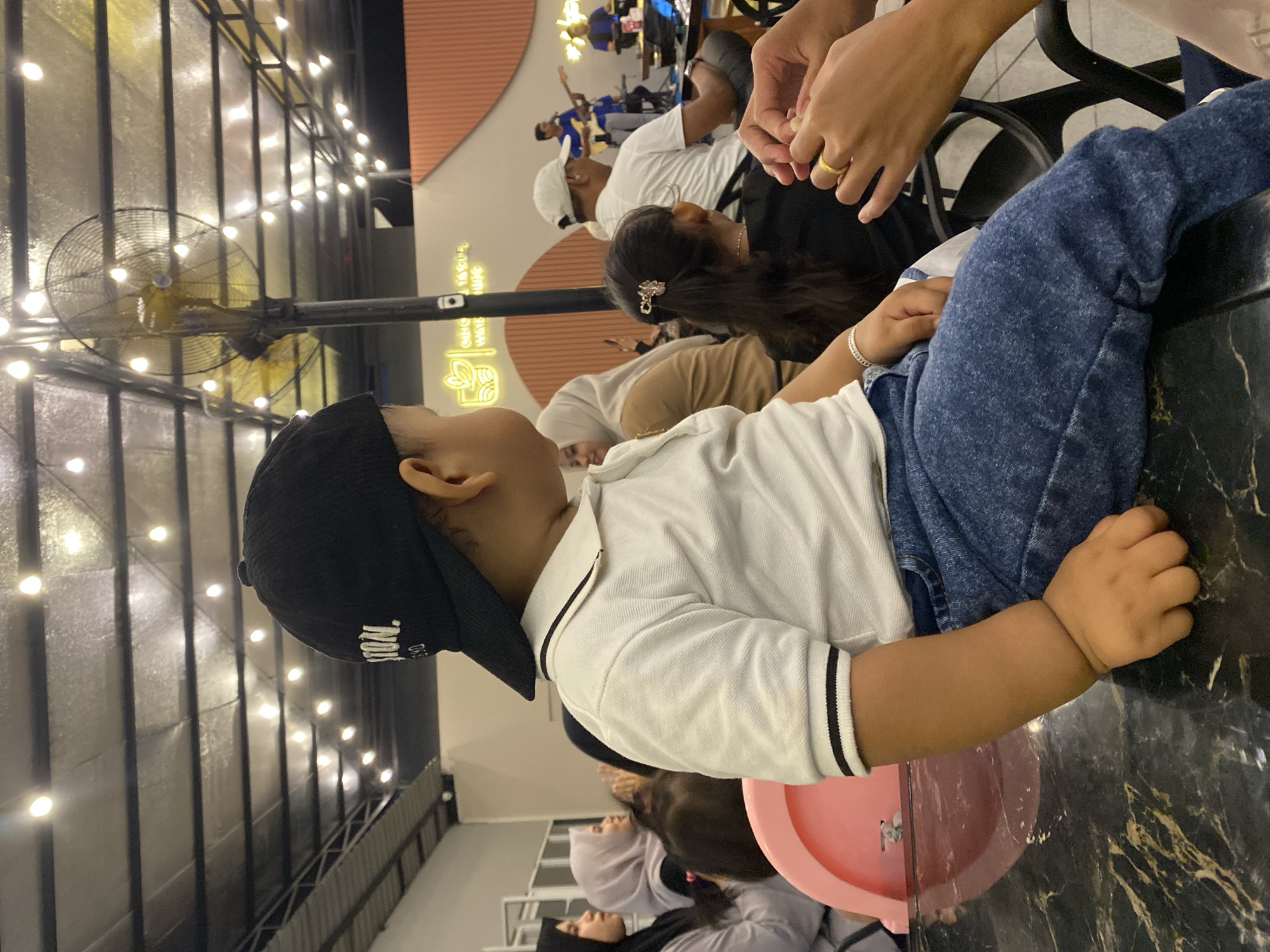 A sideways-oriented medium shot shows a child wearing a white collared shirt with black trim, blue jeans, and a black baseball cap, sitting in a pink high chair at a table in a well-lit indoor setting. The table has a dark, marbled surface. Other people are visible in the background, including a woman with dark hair and a hair clip and a person wearing a white shirt. There are hands visible in the upper right corner. Above the scene, there are hanging lights and industrial fans. The scene appears to be in a restaurant or indoor dining area, possibly in Lhokseumawe, Indonesia.