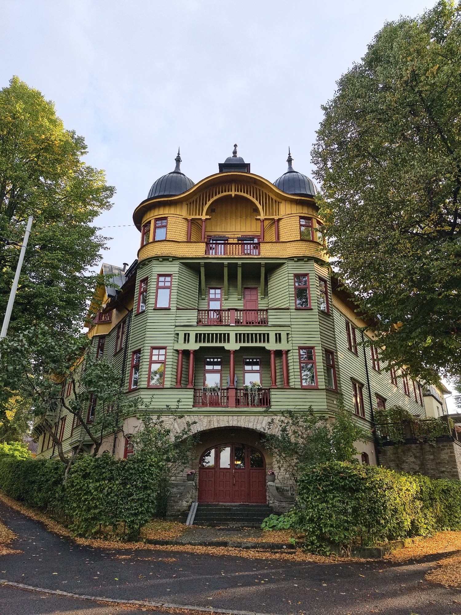 A multi-story building with a distinctive green wooden exterior and yellow trim is the central subject of the image, viewed from a low angle. The building has a curved facade and is topped with dark domed turrets. Red-framed windows are visible throughout the structure, along with several balconies with red railings. The building is surrounded by mature trees with green and yellowing foliage, suggesting an autumn setting. In the foreground, a paved pathway curves around a dense green hedge, and fallen orange leaves are scattered on the ground. The entrance to the building features an arched stone facade with large red double doors and a set of stairs leading up to it. The sky is overcast with patches of blue visible. The building is identified as being located in the city of Norrköping, Sweden. There are no people or discernible actions occurring in the image.