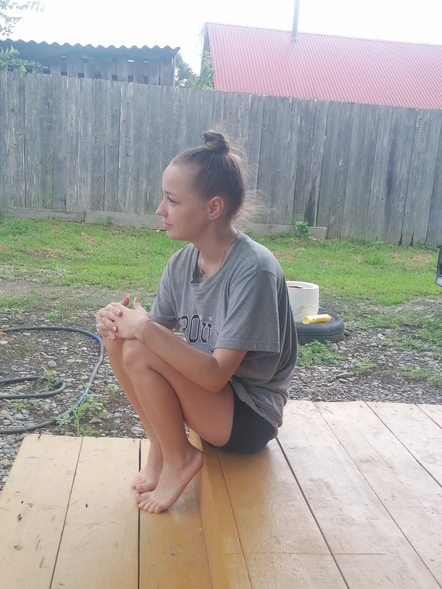 A young Caucasian female with her hair in a bun is sitting in a crouched position on a wooden porch. She is wearing a grey t-shirt and black shorts. Her hands are clasped in her lap. The background consists of a weathered wooden fence, some grass, and a red-roofed building. The scene appears to be in a rural or suburban area, possibly Novopokrovka, Russia, given the context.