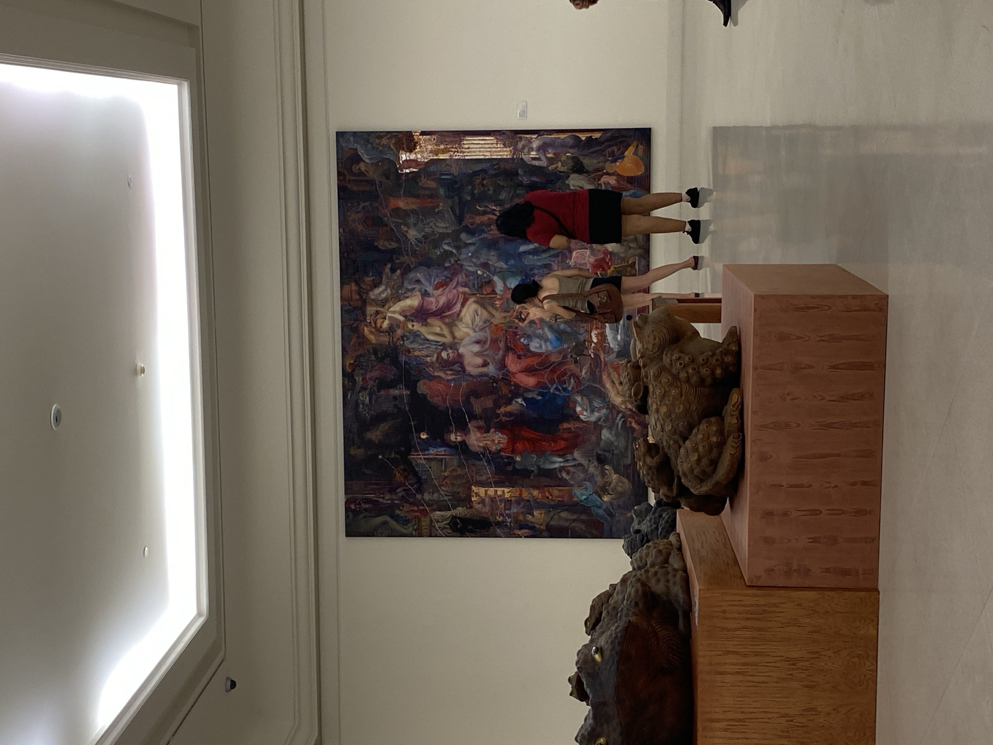 The image depicts an interior scene likely in a museum or art gallery in Bangkok, Thailand. A large painting dominates the scene, hanging on a wall. Two individuals are standing in front of the painting, seemingly observing it. One person is wearing a red shirt and a black skirt, while the other wears a light-colored top and dark shorts, carrying a brown bag. In the foreground is a display stand with sculpted figures, possibly depictions of lions or other mythological creatures, adding to the ambiance of a cultural or artistic space. The left side of the image shows a brightly lit white wall.