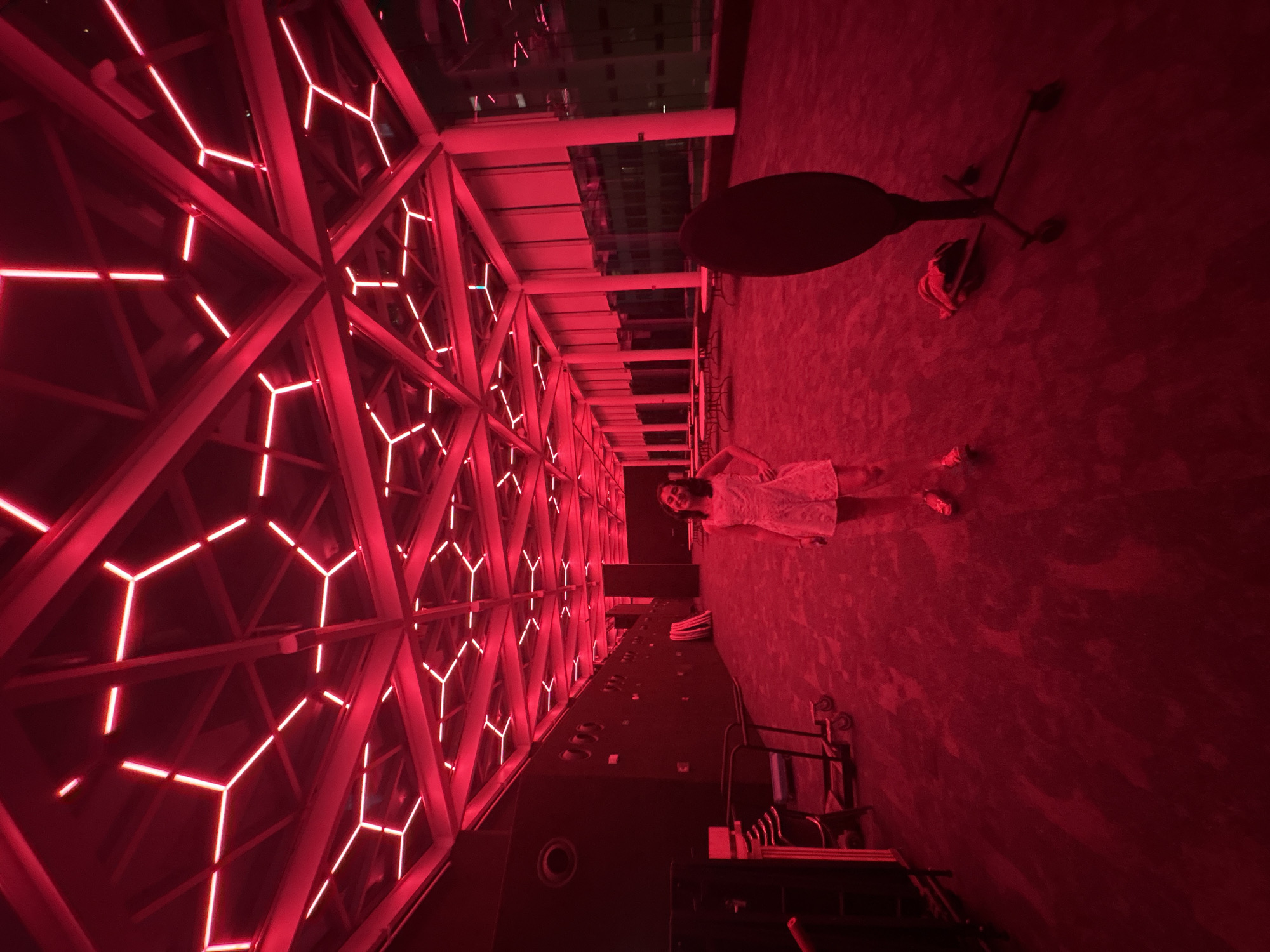 A sideways, full shot shows a woman in a white dress standing on a red-lit floor. The space has red-lit geometrically patterned windows and a table and lamp behind the woman. The location is likely in Mexico City, Mexico, due to the architectural style resembling structures in that city. There is a chair and what looks like a stand below the woman.