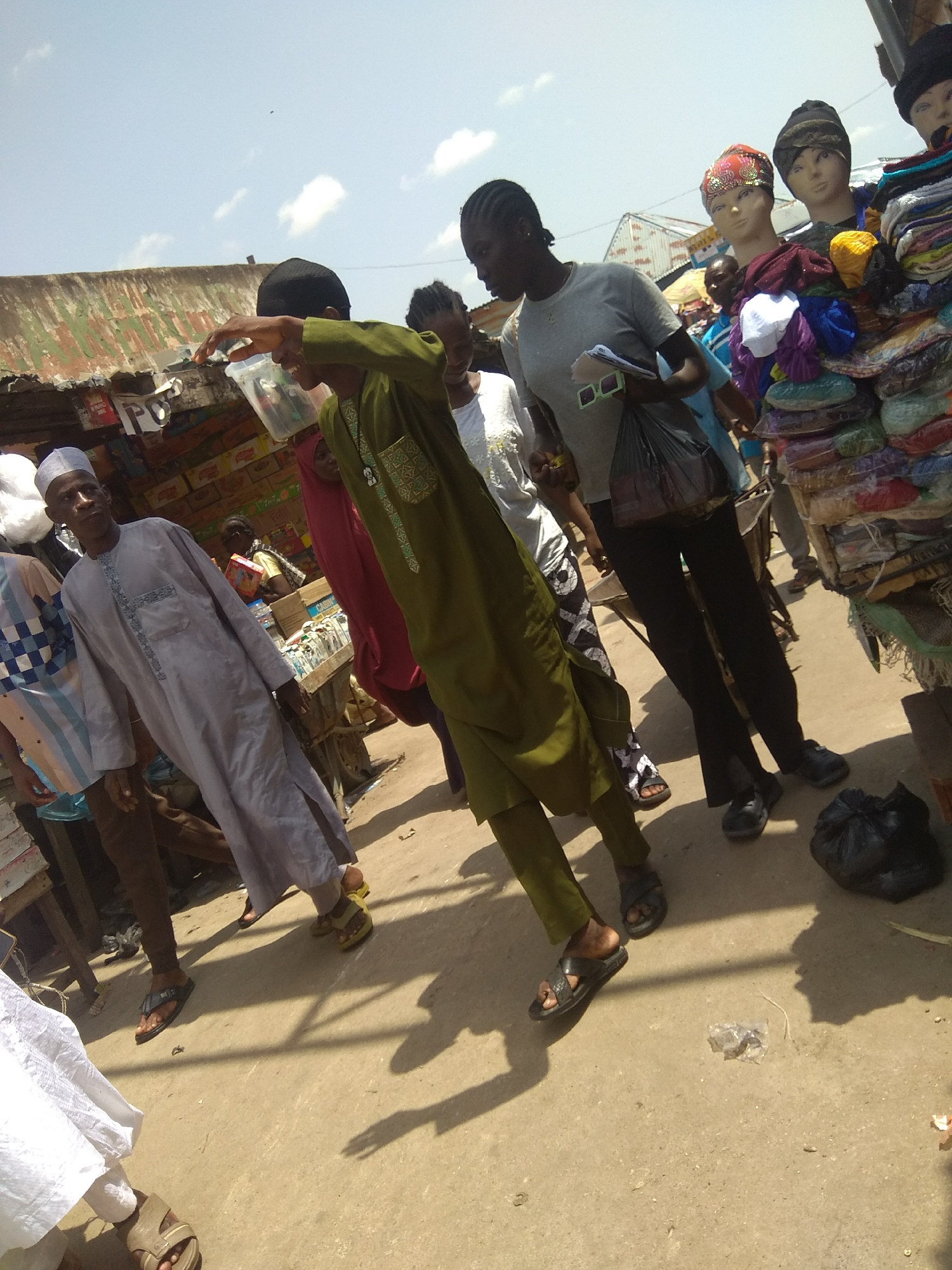 This media file captures a bustling outdoor market scene, likely in Jalingo, Nigeria. The image is dominated by several individuals engaged in everyday activities. In the foreground, a young person in a green robe is walking while holding up a clear plastic cup, their expression suggesting curiosity or interest. To their left, an older man in a light grey traditional outfit and a white cap stands with his hands clasped. Behind him, another person in a blue and white checkered shirt is partially visible.
Further back, a woman in a grey t-shirt and black pants walks with a shopping bag and what appears to be sunglasses in her hand. To her right, a display of colorful headscarves and other textiles is prominently featured, with mannequins displaying some of the items. The background reveals market stalls with corrugated metal roofs, suggesting a permanent or semi-permanent marketplace.
The time of day appears to be daytime, with bright sunlight casting distinct shadows on the dusty ground, indicating fair weather. The overall atmosphere is one of activity and commerce. While no explicit text is clearly legible, some signs are visible on the market stalls, suggesting product labels or vendor information. The presence of traditional attire and the general market setting strongly point to a location within Nigeria.
