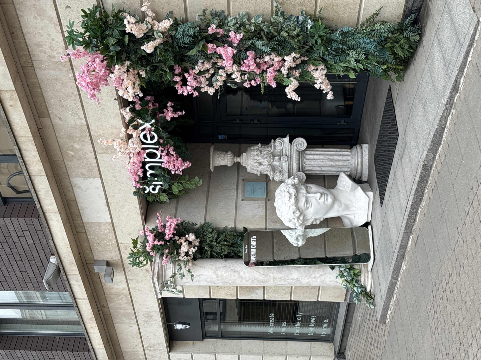 The image captures the elaborately decorated entrance of a building, likely a commercial establishment, under daytime conditions. The setting appears to be an urban environment, with paved sidewalks visible at the bottom of the frame.
The entrance features dark-framed double doors adorned with an archway of artificial greenery and delicate pink and white faux flowers. Below the floral arch, a white, vertically oriented illuminated sign spells out "Šimplex". On the left side, a window displays the text: "We create smiles for the city that loves to smile."
To the right of the entrance, decorative white classical-style elements are prominent. These include a fluted column capital with volutes and a smaller vase-like ornament. A large white bust of a classical male figure, resembling ancient Greek or Roman sculpture, rests on a pedestal to the right. In front of the bust stands a rectangular mirror, reflecting parts of the sculpture and the greenery. A black sign with white text, "ВРЕМЯ СИЯТЬ" (VREMYA SIYAT', meaning "TIME TO SHINE" in Russian), is placed near the mirror.
The building's facade is constructed from light-colored, possibly stone or concrete, panels. A dark grey mat is positioned in front of the doors. To the far left, another building section with reddish-brown brick-like patterns and windows is partially visible. The scene suggests a business that combines classical aesthetics with modern branding to create an inviting and potentially sophisticated atmosphere.