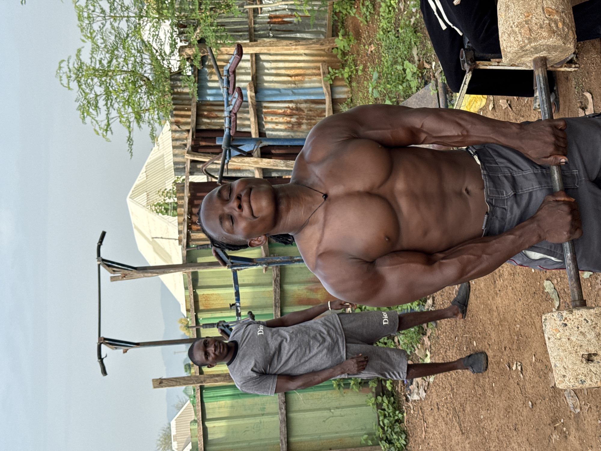 Two men are engaged in a weightlifting exercise in an outdoor gym in Akwanga, Nigeria. The man in the foreground, shirtless and muscular, is holding a barbell with what appears to be a homemade weight. He is wearing grey shorts and has his eyes closed with a determined expression. In the background, a second man, wearing a grey t-shirt and shorts, stands watching. The gym is set up with makeshift equipment, including a pull-up bar made of metal poles and wood. The surroundings include corrugated metal fencing, lush green vegetation, and a cloudy sky, suggesting an overcast day. The ground is dirt and gravel. The scene captures a moment of intense physical exertion in a rustic, community-based fitness setting.