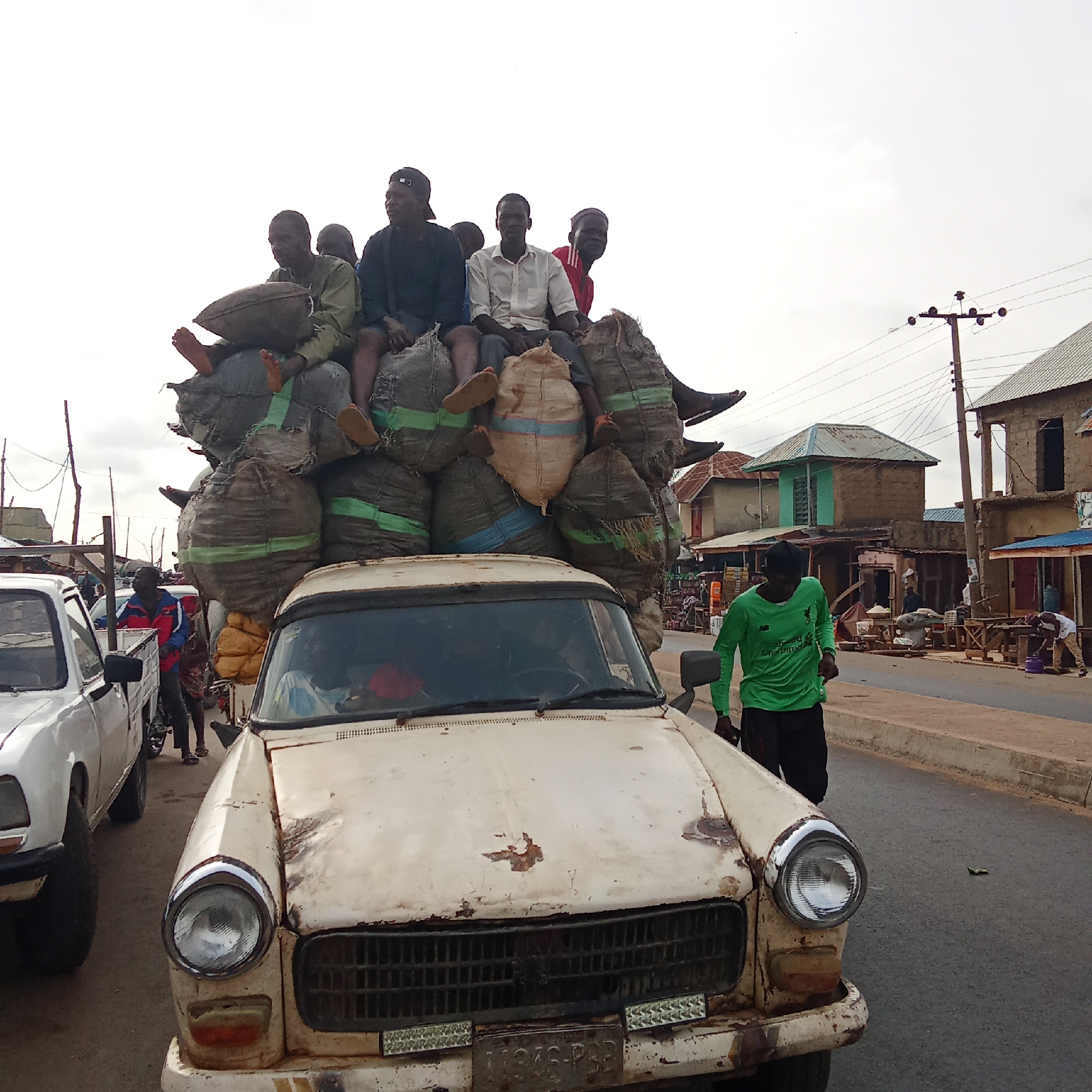 In Akwanga, Nigeria, an old, white car is heavily laden with large sacks on its roof. Several people are sitting atop the sacks. A man in a green shirt stands beside the car. Another white pickup truck is to the left of the car. Buildings and shops line the street in the background. An electrical pole stands near one of the buildings.