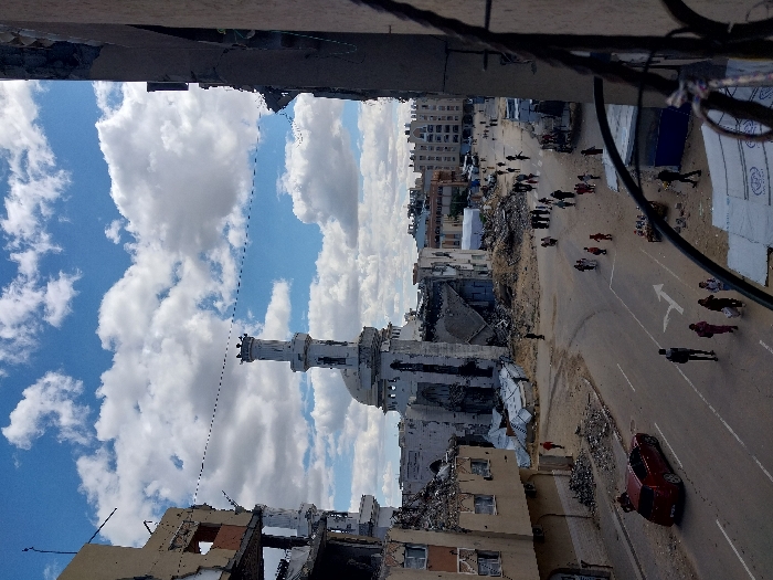 The image depicts a wide-angle street scene in Khan Yunis, Palestinian Territory, under a sky with scattered white cumulus clouds against a blue background. The scene is characterized by extensive urban damage.
In the midground, a prominent mosque stands severely damaged, with much of its main structure reduced to rubble. Its minaret remains largely intact, capped with a dome, positioned left of center. To the immediate left of the mosque, a multi-story building exhibits catastrophic structural failure, with walls collapsed, exposing interior floors and rebar. Piles of concrete rubble, twisted metal, and debris are scattered across a wide area surrounding these structures and extending into what appears to be an open dirt lot.
A paved road curves through the foreground and midground. On the left side of this road, a red sedan is either parked or stationary. Numerous individuals, including adults and children, are visible walking on the road and traversing the dirt areas. A group of approximately five adults and several children walk away from the camera in the central foreground on the road, where a white right-turn arrow and a solid white line are painted. Other individuals are dispersed further down the road and within the debris-laden areas.
To the far left, a severely damaged multi-story building, likely the vantage point, shows exposed internal elements. On the far right, a wall section of a building, also damaged, partially frames the scene. A thin, dark line or wire spans horizontally across the upper midground sky, connecting structures on the left and right sides of the frame. In the background, further down the main thoroughfare, more buildings are visible, some appearing less damaged, alongside a large white tent-like structure.