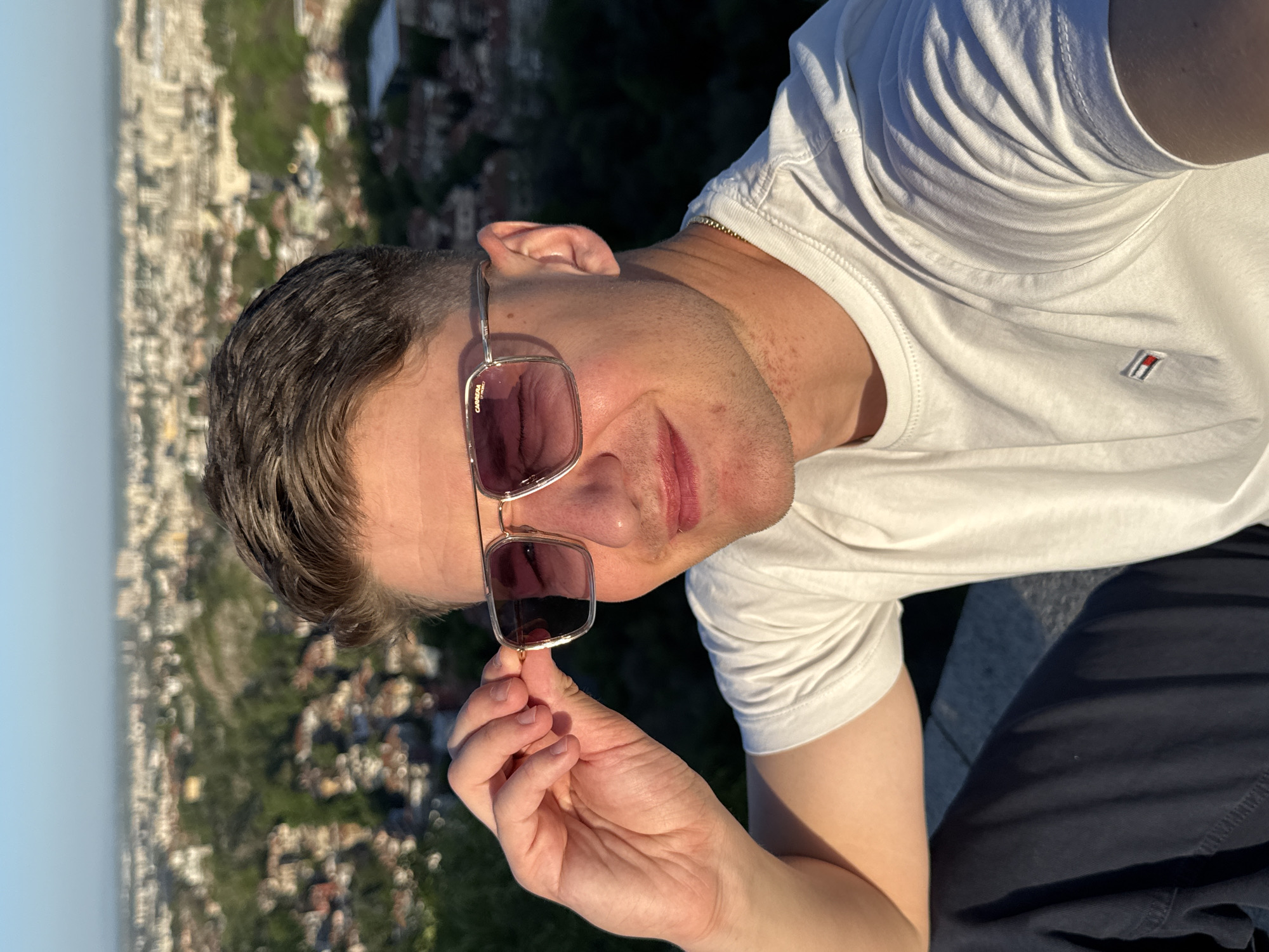 A man, likely in his twenties or thirties, is featured in a sunlit selfie, taken from an elevated outdoor perspective overlooking a city. He has short, dark hair and is wearing a white short-sleeved t-shirt with dark blue trousers. A gold chain is visible around his neck.
He is wearing stylish square-framed sunglasses with dark, possibly purple-tinted lenses. He gently holds the right temple of the sunglasses with his right hand, pulling them down slightly to reveal part of his face and a subtle smile. The brand name "CARRERA" and "BY SA..." (likely Safilo) are visible on the side of the sunglasses frame. On the left chest of his white t-shirt, there is a small rectangular logo featuring horizontal stripes in red, white, and blue.
The background reveals a panoramic view of a sprawling cityscape, characterized by numerous buildings and patches of green, extending towards a distant, hazy horizon under a clear, bright sky. The warm, golden light suggests the late afternoon or early evening. This scenic overlook is situated in Plovdiv, Bulgaria, with its distinctive urban landscape spread across hills. The man appears relaxed, enjoying the view and the clear weather.