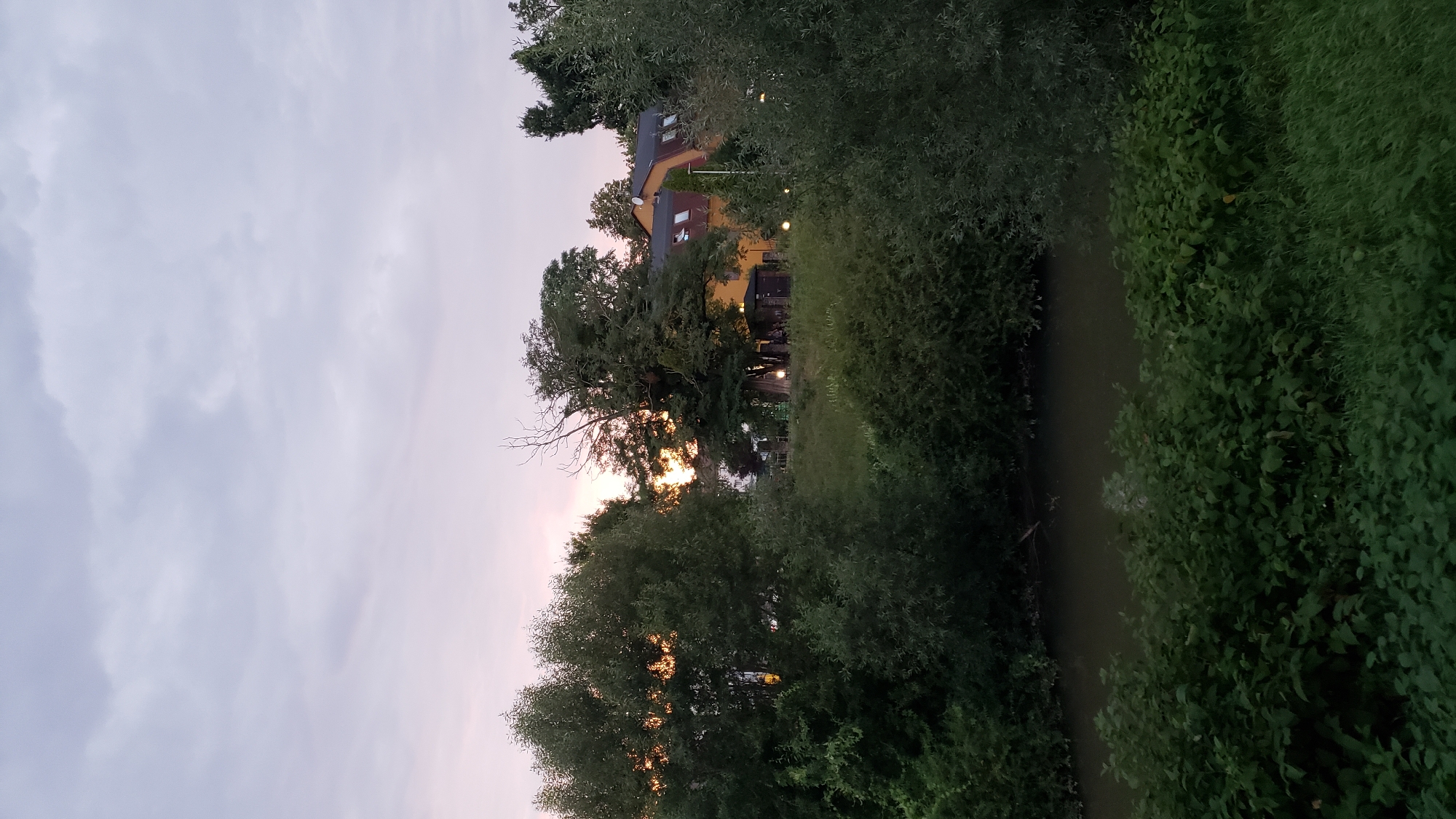 The image depicts a twilight scene with a body of water in the foreground, bordered by lush green vegetation. Beyond the water and vegetation, a cluster of trees stands on a slightly elevated bank. Behind the trees, a multi-story building with a distinctive orange roof and yellow facade is visible. The sky is overcast with clouds, tinged with the soft colors of sunset. The overall atmosphere is tranquil and natural, suggesting a park or riverside area. This scene is consistent with locations such as the park surrounding the Torysa river in Prešov, Slovakia, where natural landscapes often integrate with urban structures.