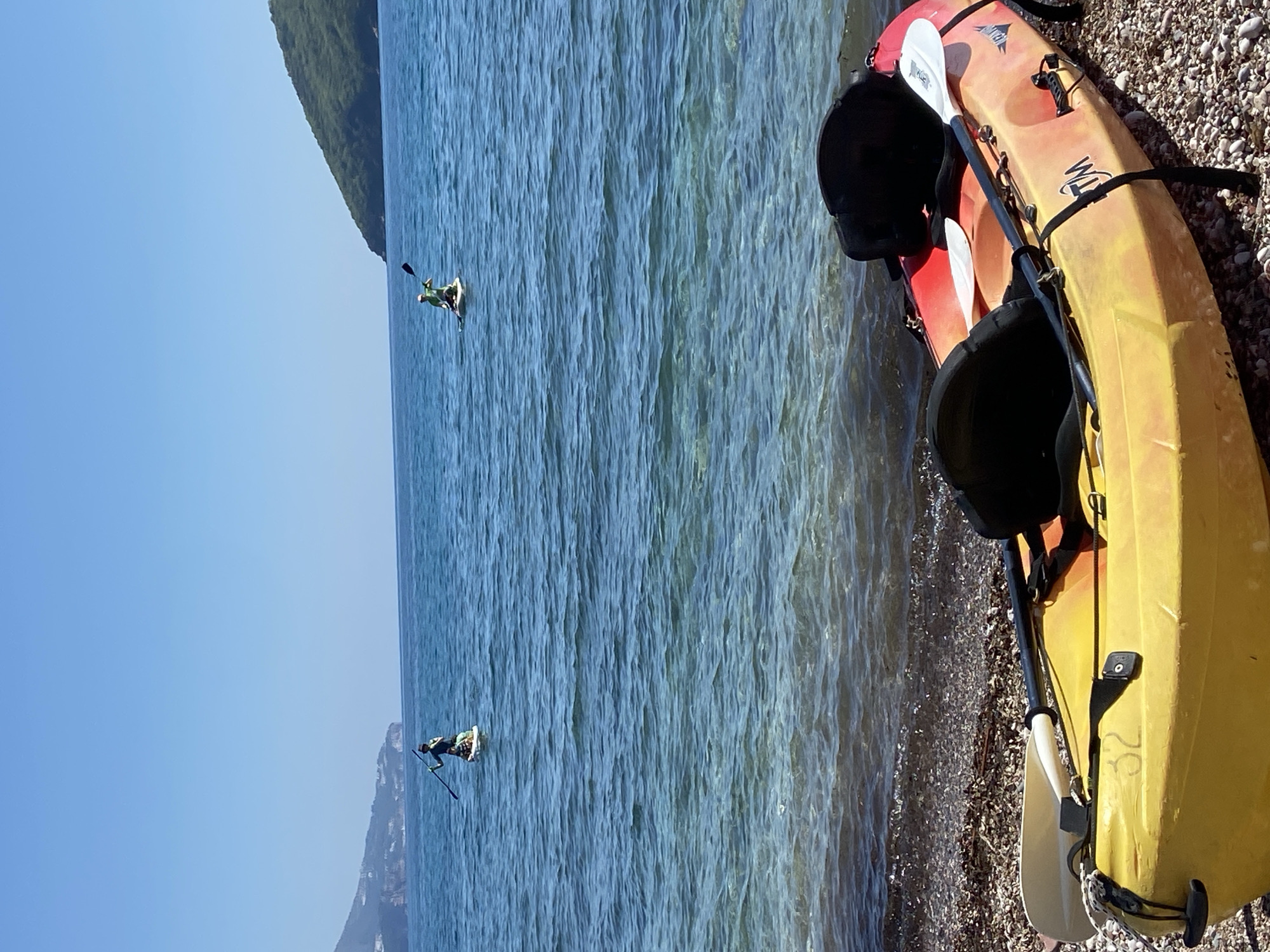 The image shows a beach scene, likely in Budva, Montenegro, with two kayaks on a pebble beach at the edge of the water. One kayak is yellow and red, with two black seat attachments and paddles. A second kayak is partially visible behind it. Two people are paddling in the calm, blue water, one closer to the foreground and the other near a headland in the background. The sky is clear and blue. The headlands are covered with trees.