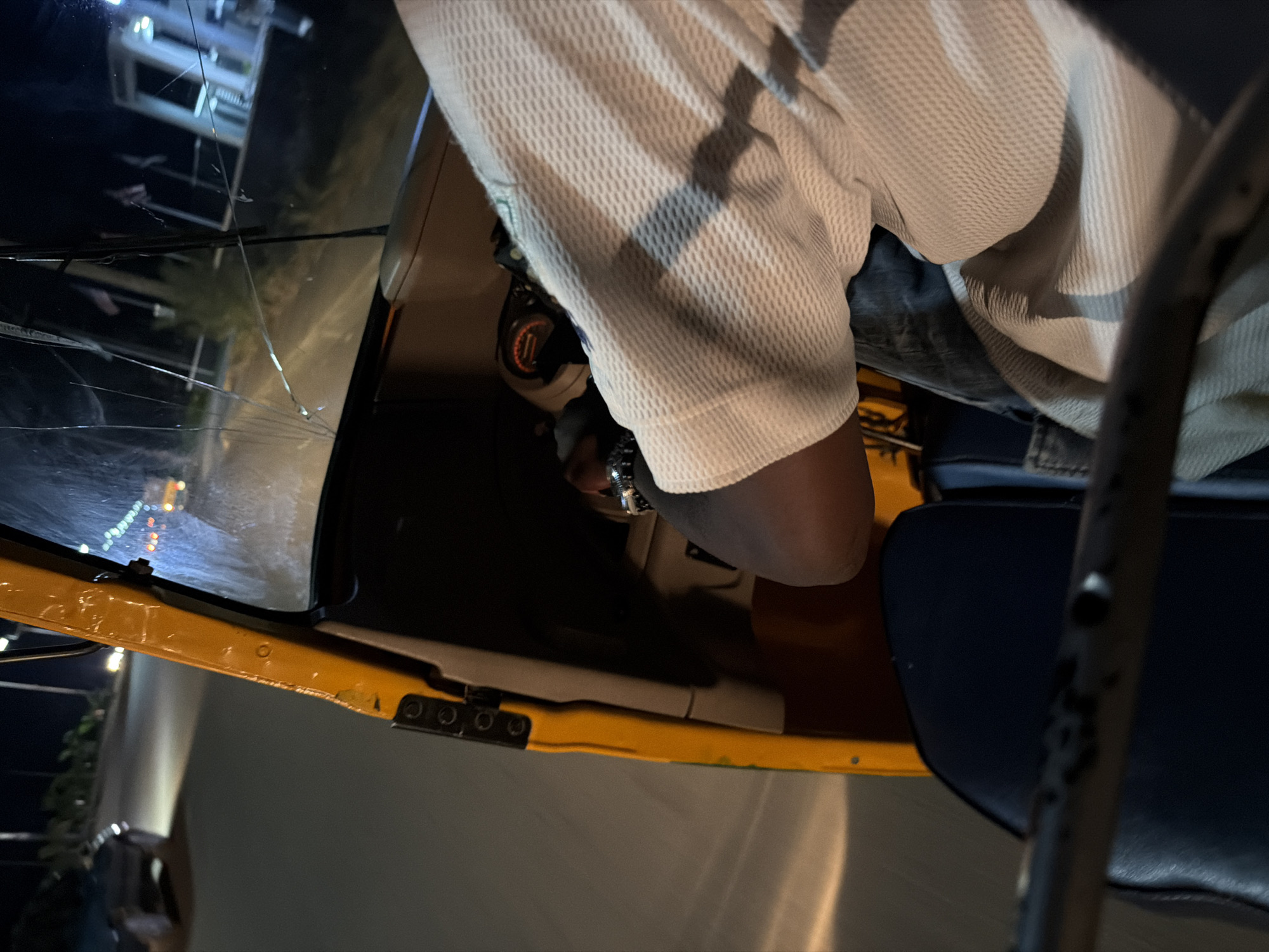 A nighttime view from inside a yellow auto-rickshaw in Bukuru, Nigeria, shows the driver's arm and shoulder wearing a light-colored, textured shirt. The driver's left hand is visible gripping the steering wheel, which has a circular gauge with orange markings. The windshield of the auto-rickshaw is cracked. The road ahead is illuminated by headlights, and other vehicles and streetlights are visible in the distance. The scene suggests the auto-rickshaw is in motion.
