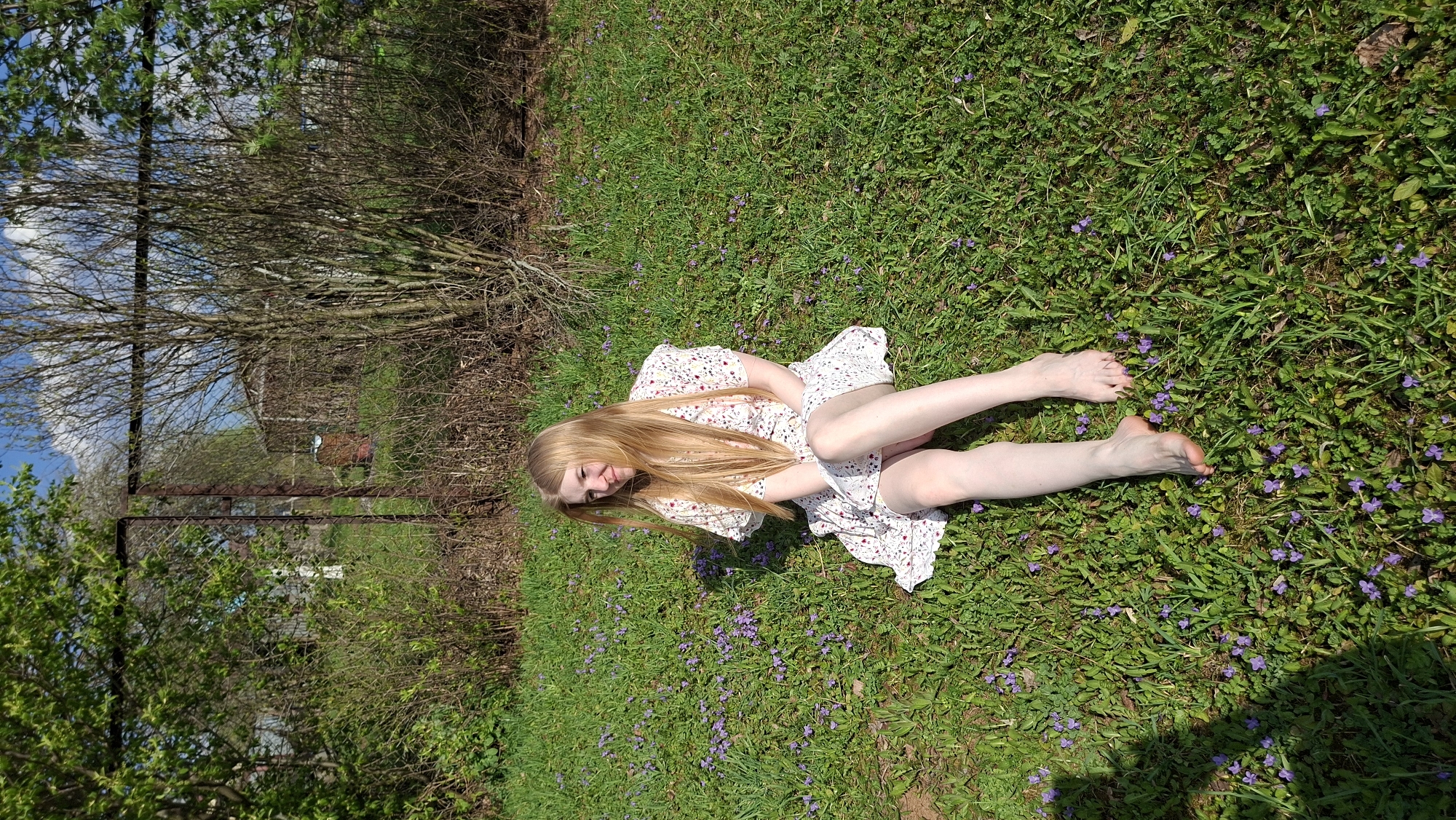 A fair-skinned young woman with long blonde hair is lying on her side in a grassy field. She is wearing a light-colored floral print dress. The field is dotted with small purple flowers. In the background, there are trees, bushes, a utility pole, and a fence. The sky is visible through the trees. The location is Большой Букор, Russia.