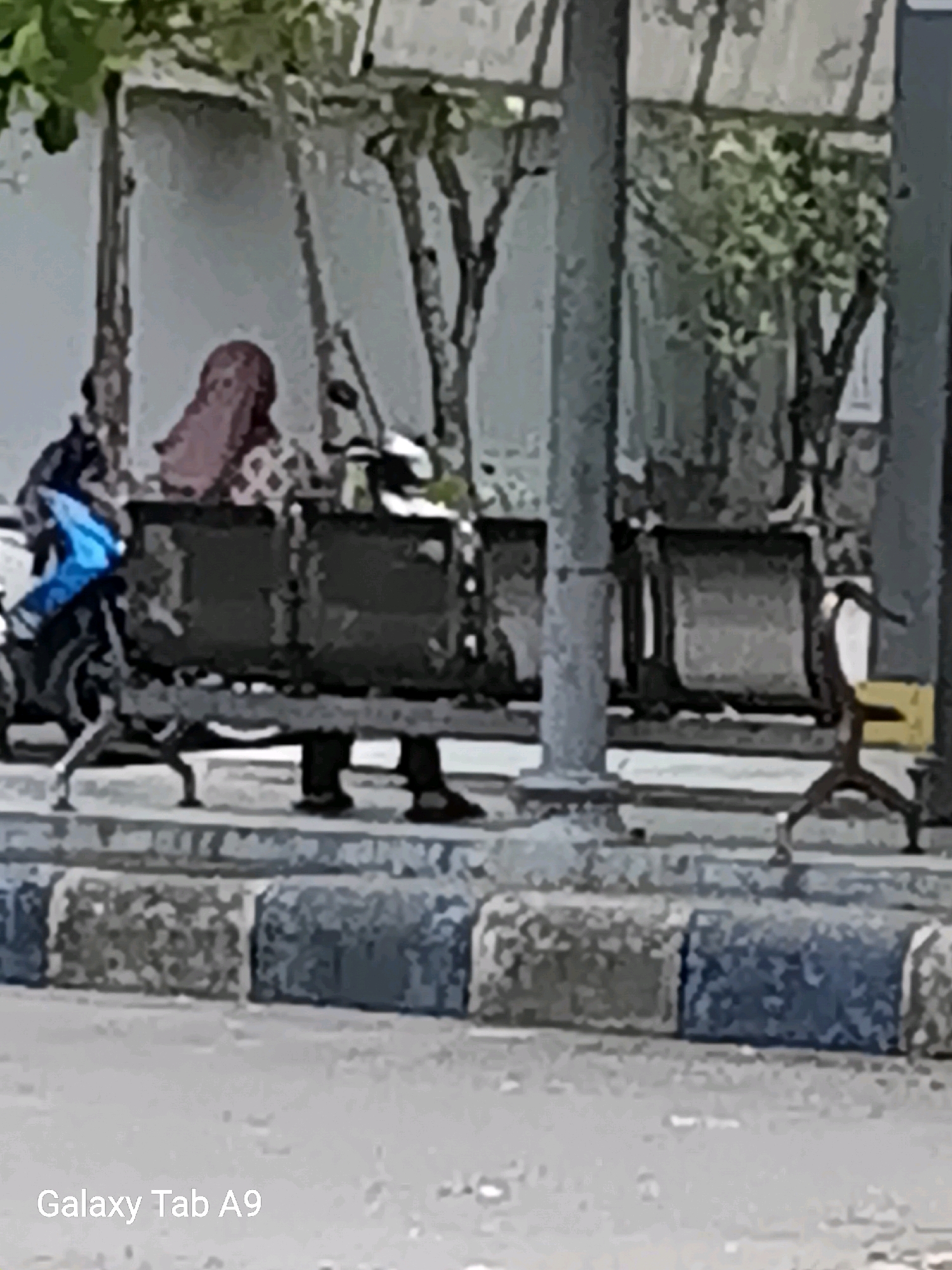 A person wearing a headscarf and patterned clothing sits on a bench under a shelter. Next to them, a person wearing a blue garment is partially visible, as is a motorcycle. Another person is seen standing to the right of the bench, holding a yellow object. The bench is made of dark material and is supported by legs. The surrounding area appears to be a public space with trees and a textured ground. The image includes the text "Galaxy Tab A9" in the lower left corner. The scene suggests a waiting area, possibly at a bus stop or public transport hub.