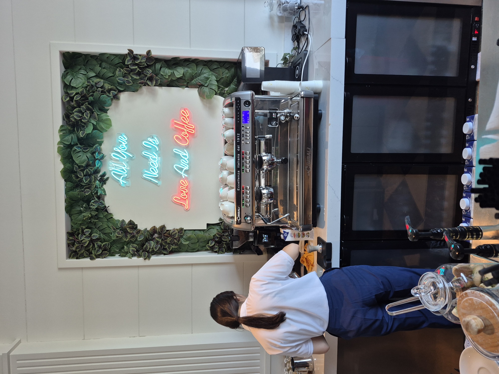 The image shows a person wearing a white t-shirt and blue pants standing behind a coffee machine. The coffee machine is a La Cimbali model. Behind the coffee machine is a sign that reads "All You Need Is Love and Coffee". Above the coffee machine are glass jars. The background is a white wall. The setting appears to be a cafe or coffee shop, potentially located in Alanya, Turkey.