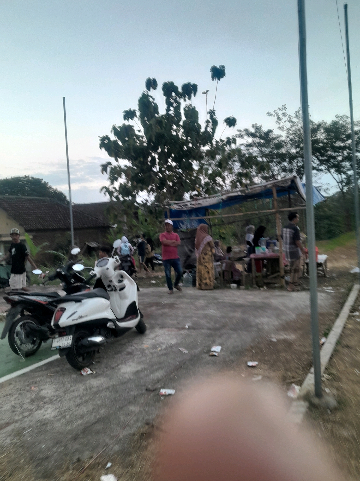 In Genteng, Indonesia, a group of people are gathered near a roadside stall. Two motorcycles are parked prominently in the foreground, one white and one black. The white scooter has a license plate that reads "P 4397 CBX". Behind the motorcycles, a stall with a blue tarp roof is set up, with several individuals standing around it. One person in a pink shirt and white cap is standing to the right, facing away from the camera. Another person in a patterned dress and headscarf is visible near the stall, as is a person in a plaid shirt. A building with a tiled roof is visible in the background, along with trees and utility poles. The ground is paved and appears to be slightly wet in places, with scattered litter. The sky is overcast and light.