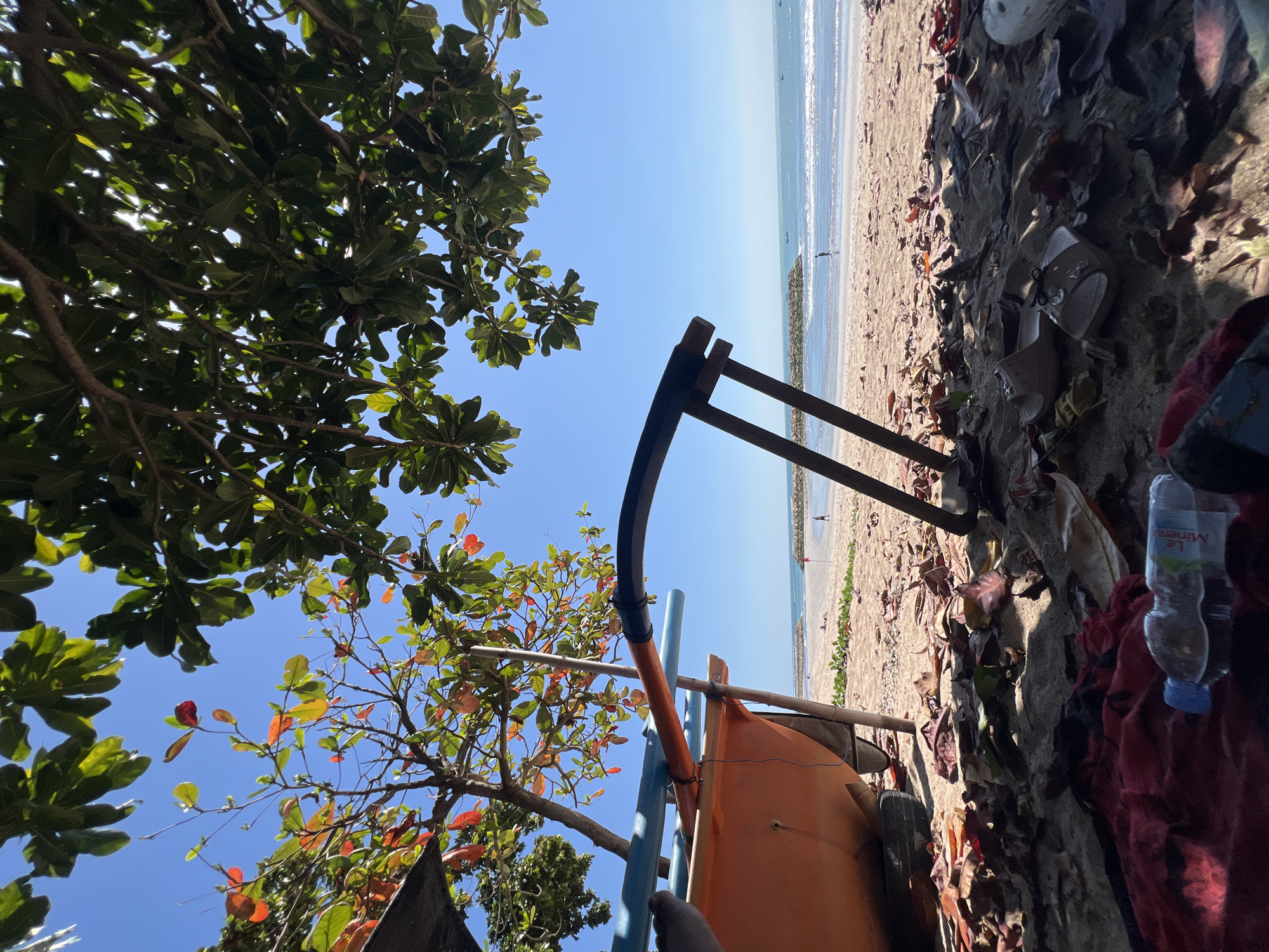 The image captures a sunny day on a beach in Kuta, Indonesia, viewed from a low-angle perspective. In the foreground, dry leaves and scattered debris cover the sandy ground. A partially visible orange boat rests on the sand, supported by wooden poles. To the left, a red cloth and a plastic water bottle are on the ground.
Beyond the foreground, the beach stretches towards the ocean. Gentle waves lap at the shore, and in the distance, there are stone breakwaters and a few small boats. The sky is clear and bright blue. Trees with green and reddish-colored leaves partially shade the scene, with their branches reaching into the frame from the top. A few distant figures can be seen walking on the beach and in the shallow water. The overall atmosphere is serene and tropical.