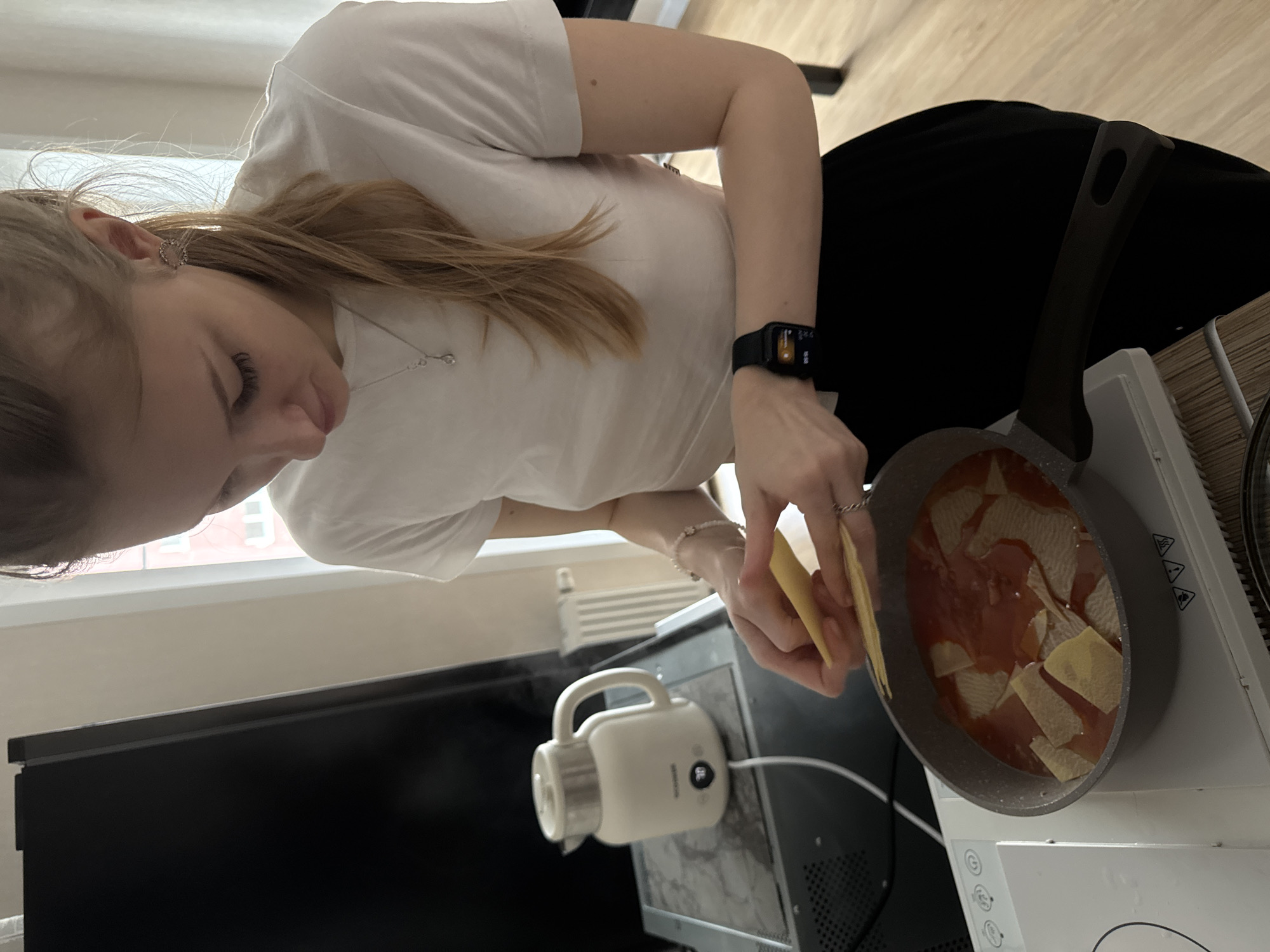 A young woman is captured in a vertical, high-angle shot, preparing food in what appears to be an indoor kitchen or cooking area in Perm, Russia. She has light brown hair, wears a white t-shirt, black pants, a silver necklace, and a smart watch on her left wrist. A silver ring is visible on her right hand.
The central activity shows her actively cooking. She is holding flat, yellow pasta sheets, possibly for lasagna, and is in the process of adding them to a grey frying pan containing a red sauce. The pan rests on a white electric or induction cooktop, which features circular control buttons with symbols for power, settings, and hazard warnings (three triangles with exclamation marks).
To the left of the cooktop, a white electric kettle or small beverage maker is plugged in and sits on a black surface, likely a microwave oven or mini-fridge. Further left, a black monitor or TV screen is visible. In the background, a large window reveals an outside building, suggesting an urban setting, with natural light illuminating the scene. A white radiator is visible below the window. The floor is light-colored wood. The woman's expression is focused on her cooking task. No specific time of day is discernible beyond daylight, and no weather details are apparent. Text on the smart watch and kettle's control panel is not clearly legible.