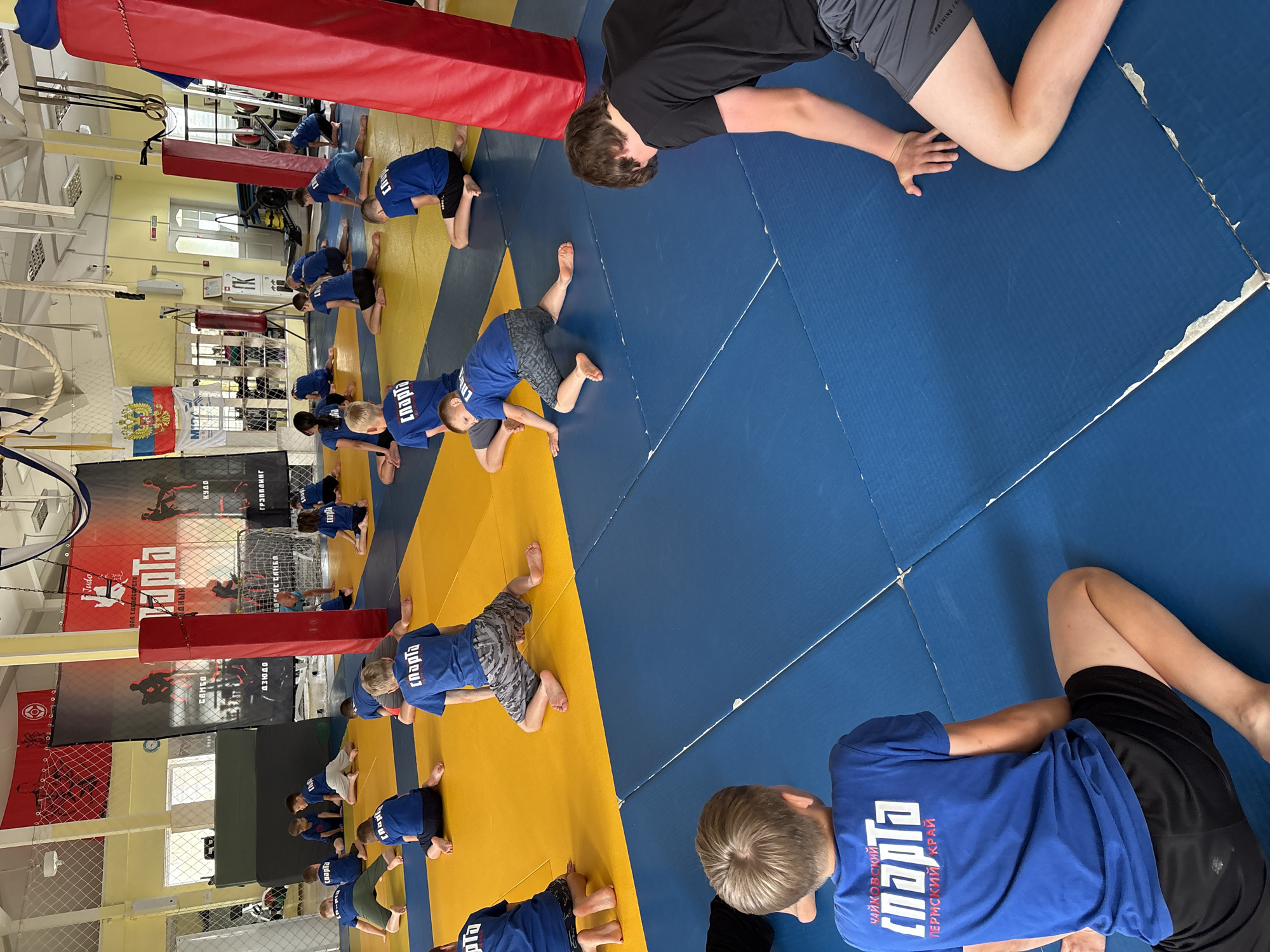 The image depicts a judo training session taking place in a gymnasium, likely located in the city of Chaikovsky, Russia, as indicated by the text on some of the participants' attire. Several young boys are engaged in stretching exercises on a blue and yellow mat. They are all wearing similar blue t-shirts with white lettering that says "СПАРТА" (Sparta) and the text "ПЕРМСКИЙ КРАЙ" (Perm Krai) below it, identifying their region. Their shorts vary in color and pattern, with some wearing camouflage and others plain black. In the background, there are judo banners with images of athletes, flags, and gym equipment, including punching bags and rings, suggesting a dedicated training facility. A large red padded pillar is prominent on the right side of the frame. The lighting appears to be artificial, common for indoor sports venues.