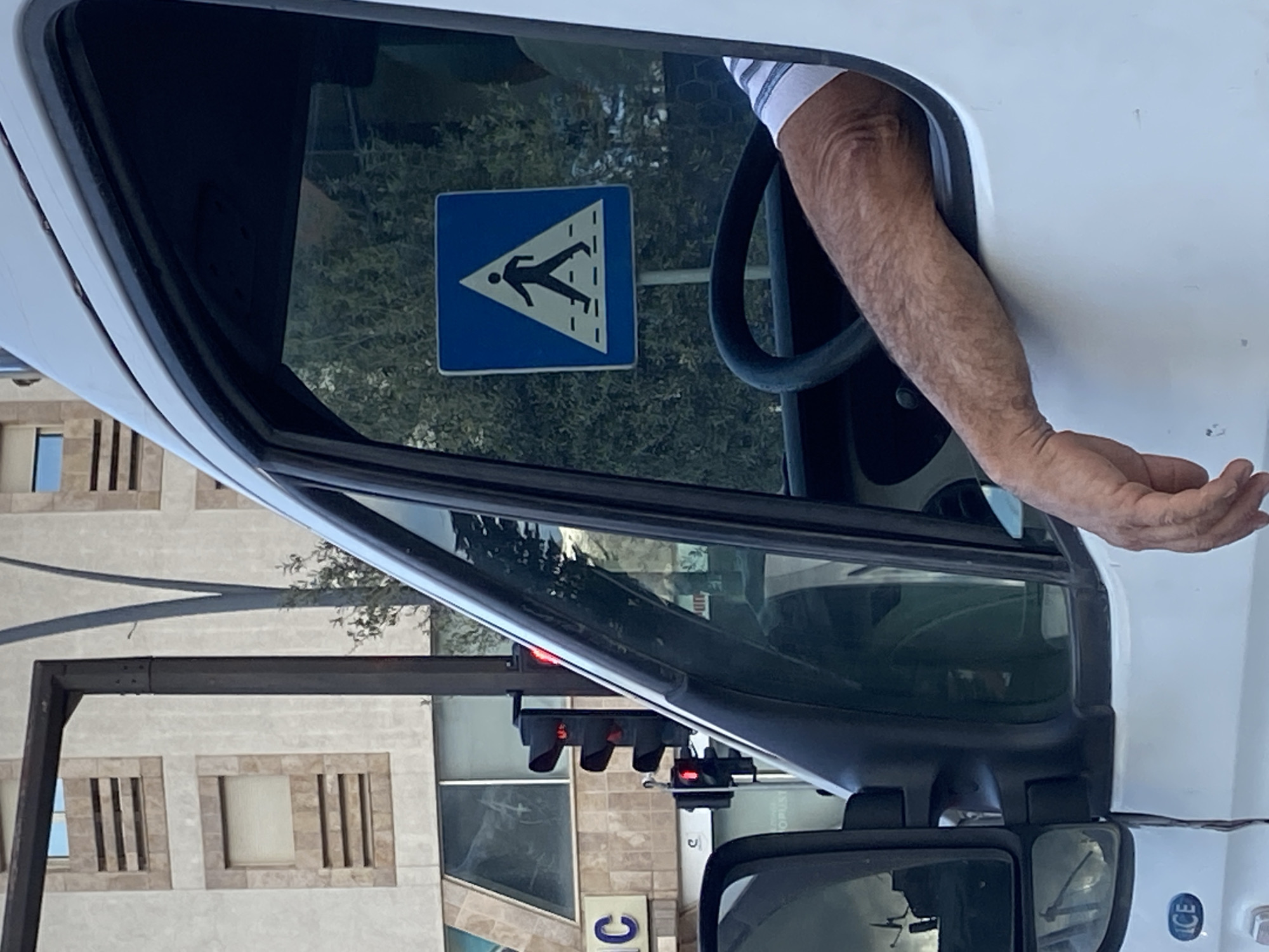 A driver's side view from inside a white car, facing forward, reveals a traffic signal with red lights, indicating a stop. A blue and white pedestrian crossing sign is visible through the passenger side window. A man's arm is extended out of the driver's side window. The background shows buildings with windows. The car is identified as "ICE" on the bottom right. The location appears to be in an urban area, possibly Budva, Montenegro.