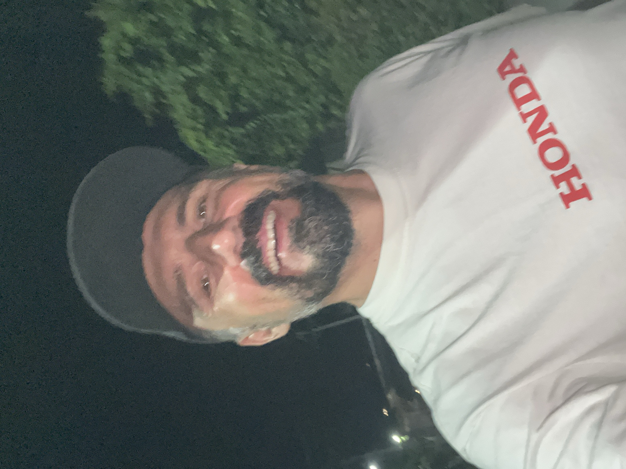 A man with a graying beard and mustache, wearing a gray baseball cap and a white t-shirt with the word "HONDA" printed vertically in red, is smiling and looking at the camera. He appears to be outdoors at night, as evidenced by the dark background and foliage partially visible. The location is บ้านบางรักษ์, Thailand.