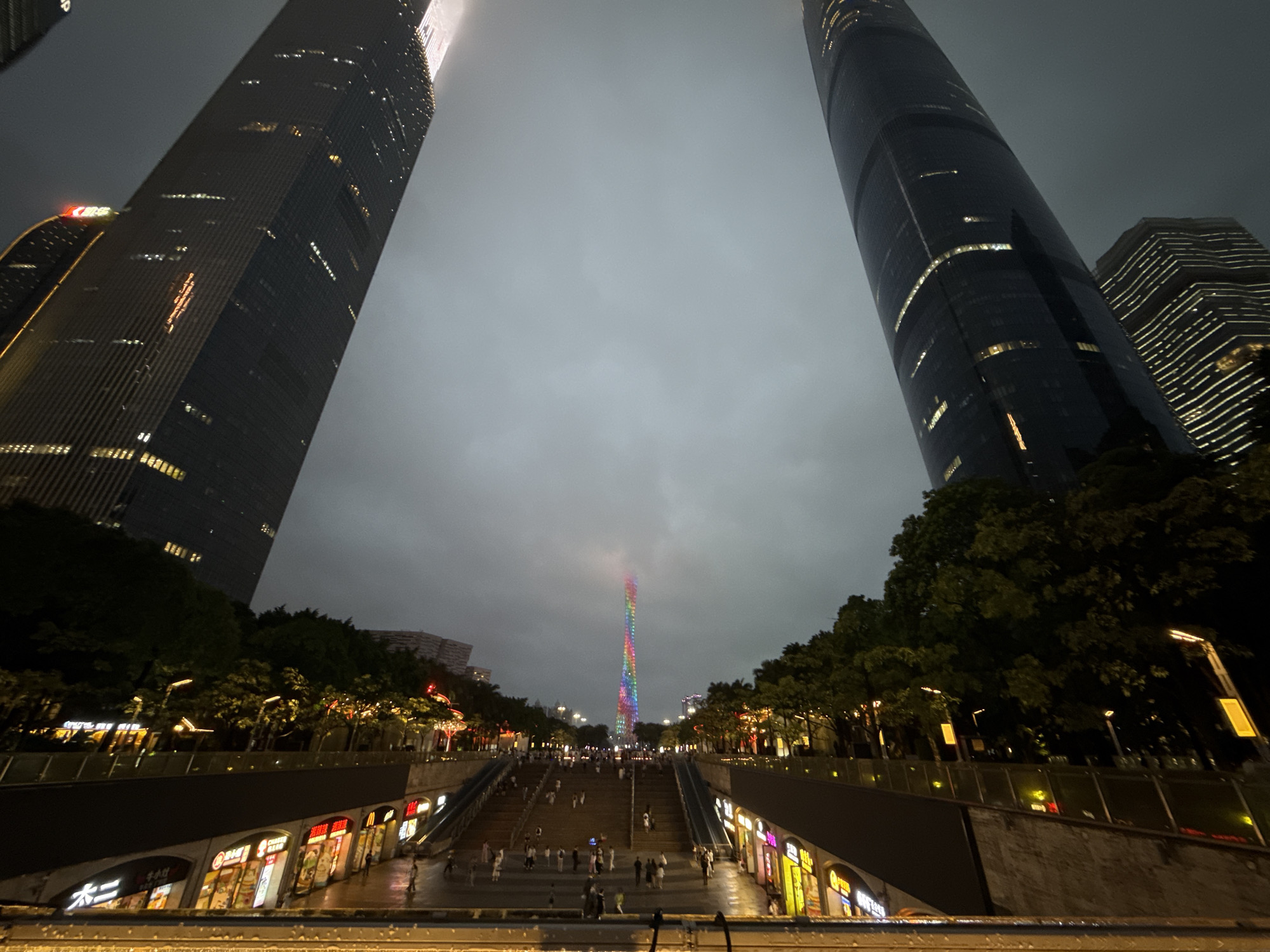 The image captures an urban scene at night in Tianhe District, China. Two skyscrapers flank a central pathway lined with trees and streetlights, leading towards the Canton Tower, illuminated in a spectrum of colors. Pedestrians are visible on the steps connecting the foreground to the base of the tower. Buildings with storefronts are on either side of the walkway, and foliage is interspersed among the built environment. The sky is overcast, creating a muted backdrop for the illuminated structures and street lighting.
