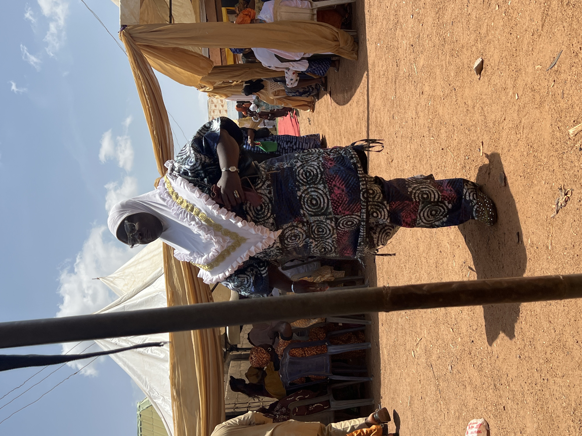 The image depicts a woman standing in an outdoor setting, presumably in Rigasa, Nigeria, based on the prompt. She is dressed in a patterned fabric, predominantly dark blue with white and red circular designs, and a white head covering. A white ruffled collar with gold trim adorns her upper garment. She wears glasses and a watch on her left wrist.
The setting appears to be an event or gathering. Other people are seated in chairs under what looks like a tent structure with draped fabric, suggesting shade provision. The ground is bare earth with some scattered debris. A section of a dark-colored horizontal pole is visible in the foreground. The sky is light blue with some clouds.