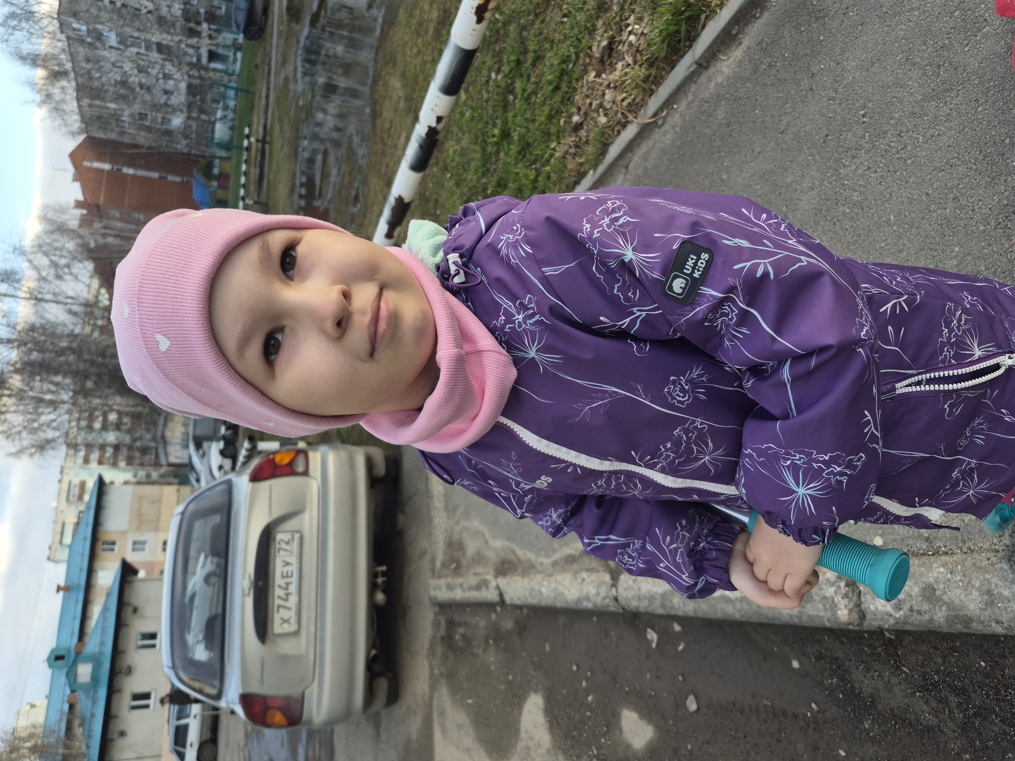 A young child wearing a pink hat and purple patterned snowsuit stands on a sidewalk next to a road in Tobolsk, Russia. The child is holding the green handle of a scooter. Behind the child, a silver car is parked on the side of the road with the license plate "X744EY72". Buildings with blue roofs are in the background, along with trees. The weather appears to be overcast.