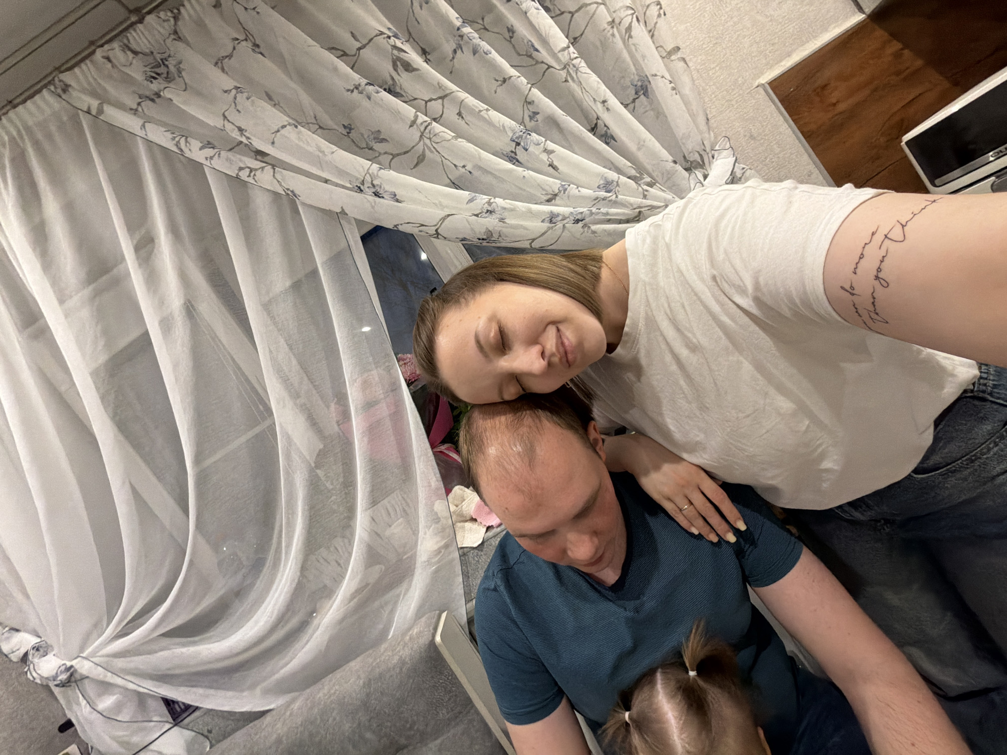 This intimate portrait captures a family moment indoors, likely in a living room or bedroom in Reshetnikovo, Russia. The scene features three individuals: a woman, a man, and a young child.
In the foreground, a woman with light brown hair, wearing a white t-shirt and blue jeans, gently rests her head on the man's head. Her eyes are closed in a peaceful smile, conveying affection and serenity. She wears a ring on her left-hand ring finger, and a visible tattoo on her right arm reads, "You are more than you think."
Below her, a man with light brown, receding hair wears a dark teal or blue ribbed t-shirt. He is looking downwards, seemingly engaged with a small child whose head, with hair tied in two pigtails, is visible in the bottom right corner of the frame. The child appears to be sitting on his lap or very close to him, suggesting a shared activity or quiet time.
The background reveals typical home furnishings. White sheer curtains, some adorned with a delicate greyish-blue floral and branch pattern, hang to the left, partially obscuring a window through which a dim, dark view suggests it might be evening or night. A grey upholstered sofa or couch is visible on the left. The wall behind the subjects is light-colored. In the upper right corner, a dark brown wooden surface, possibly a shelf, holds a small white electronic device with a black screen.
The image conveys a sense of warmth, closeness, and domestic tranquility, capturing a tender and relaxed family interaction.