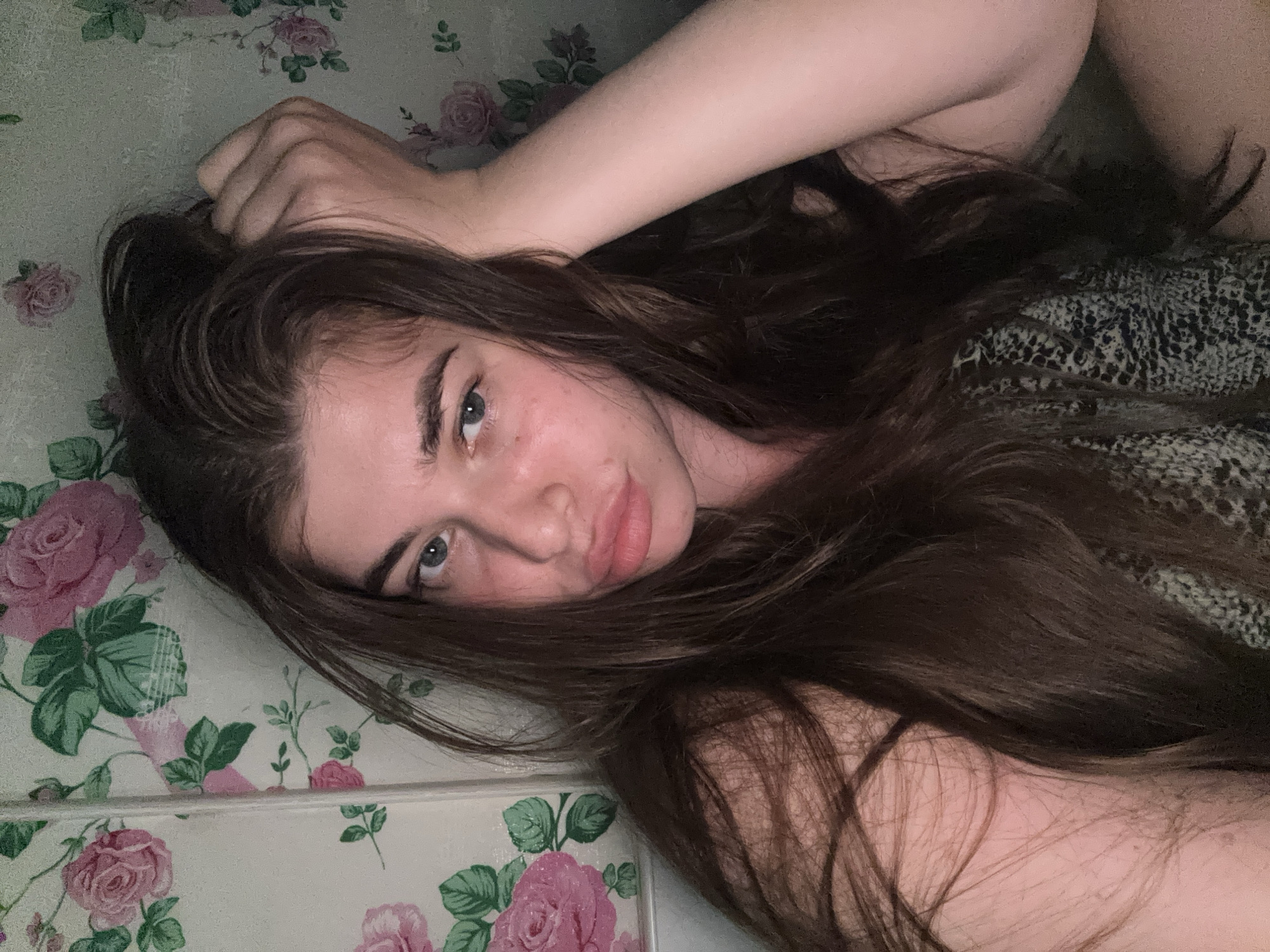 This is a headshot of a young woman with long, dark brown hair. She has blue eyes and full lips. Her right arm is raised, and her hand is resting on her head. She is wearing a patterned top. The background consists of floral wallpaper with pink roses and green leaves. The scene appears to be indoors, possibly in a bedroom or living area. There is no discernible connection to the city of Мещёры, Russia.