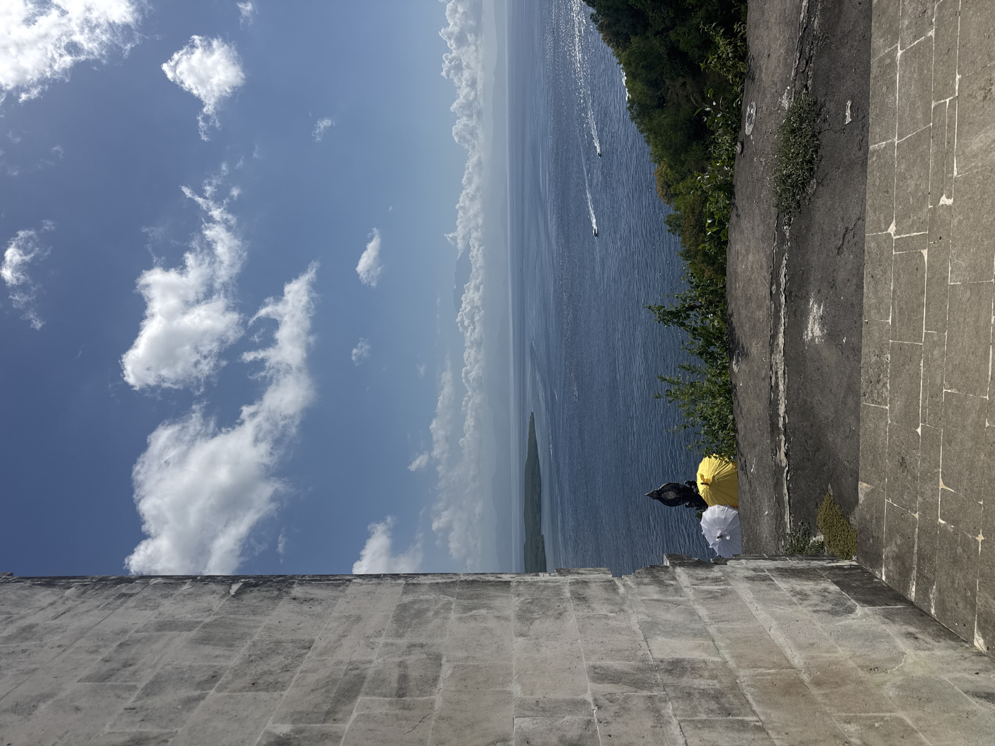 A high-angle outdoor shot captures a panoramic view of the ocean from a stone structure. In the foreground, the rough, textured stone wall of a building on the left side of the frame leads to a concrete walkway and stone steps. Two open umbrellas, one white and one yellow, are placed on the walkway near a black decorative finial. Beyond the walkway, the deep blue ocean stretches to the horizon, dotted with distant islands and a few small boats. The sky above is a vibrant blue, with scattered white clouds. Lush green trees and vegetation line the coastline in the middle ground. The overall scene suggests a scenic overlook or viewpoint in Nusa Penida, Indonesia.