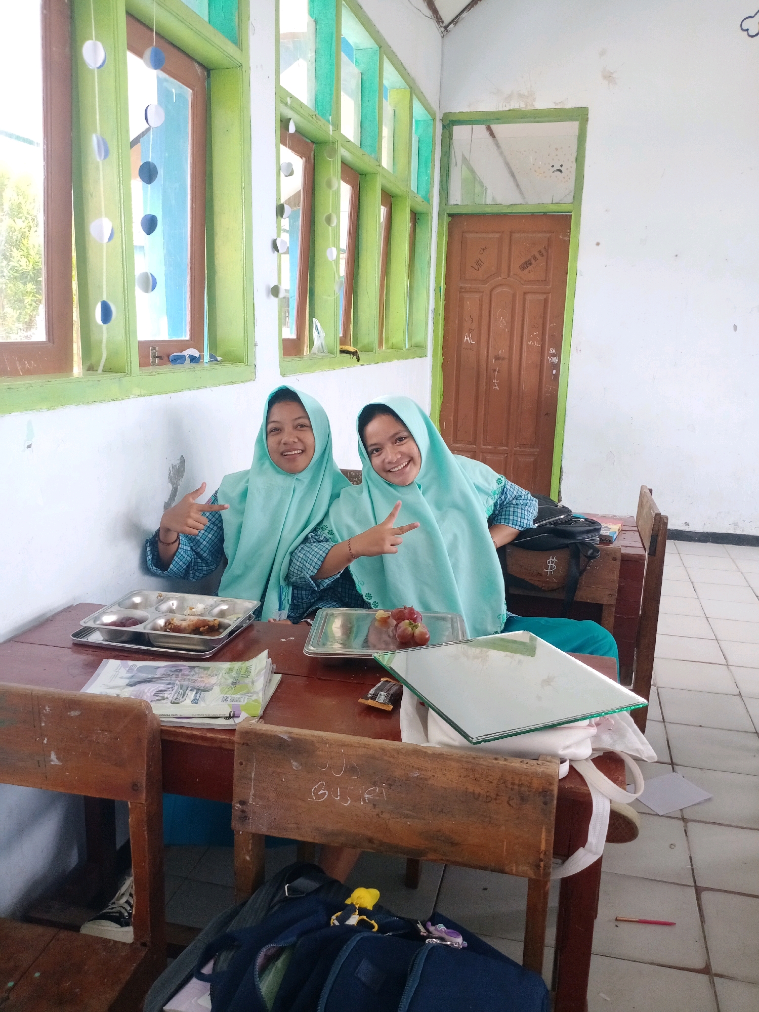 In a classroom in Tragah, Indonesia, two young girls wearing teal hijabs and checkered blue shirts are seated at a wooden desk. The girl on the left has her left hand pointing outward, while the girl on the right has her right hand in a peace sign. Both are smiling at the camera. On the desk, metal trays with food, a stack of papers, and a mirror are visible. The room has green-trimmed windows with decorations and a wooden door with graffiti. The floor is tiled. Bags and backpacks are on the floor and under the desk.