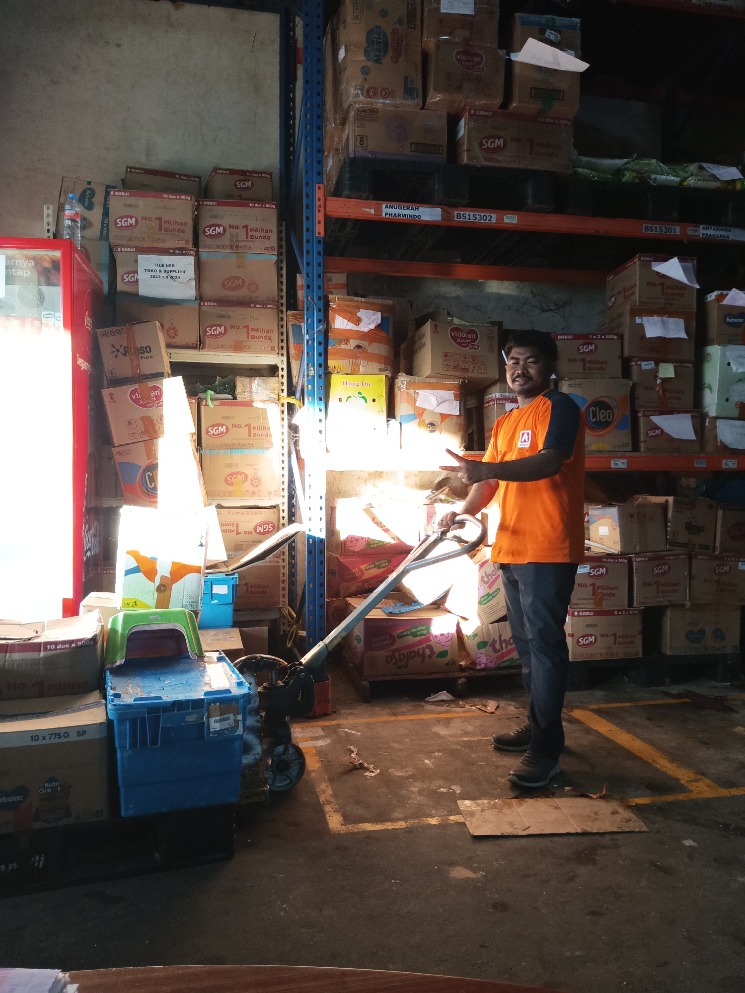 In a warehouse setting, likely in Makassar, Indonesia, a man stands holding the handle of a pallet jack. He is wearing an orange t-shirt with an emblem on the left chest and dark pants. He is positioned in front of tall shelves stacked with numerous cardboard boxes, some of which are labeled with "SGM" and "Pilihan Bunda." To his left is a red vending machine or cooler. The floor is concrete with yellow painted lines marking pathways. Various boxes, bins, and a green stool are scattered around the pallet jack and the immediate area. The lighting suggests an indoor environment with natural light filtering in from the left.