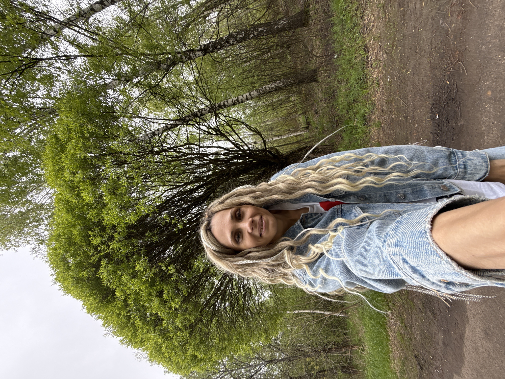 A woman with blonde hair is taking a selfie outdoors. She is wearing a light-blue denim jacket. The background shows a park with trees and a dirt path. The location is Rusanovo, Russia.