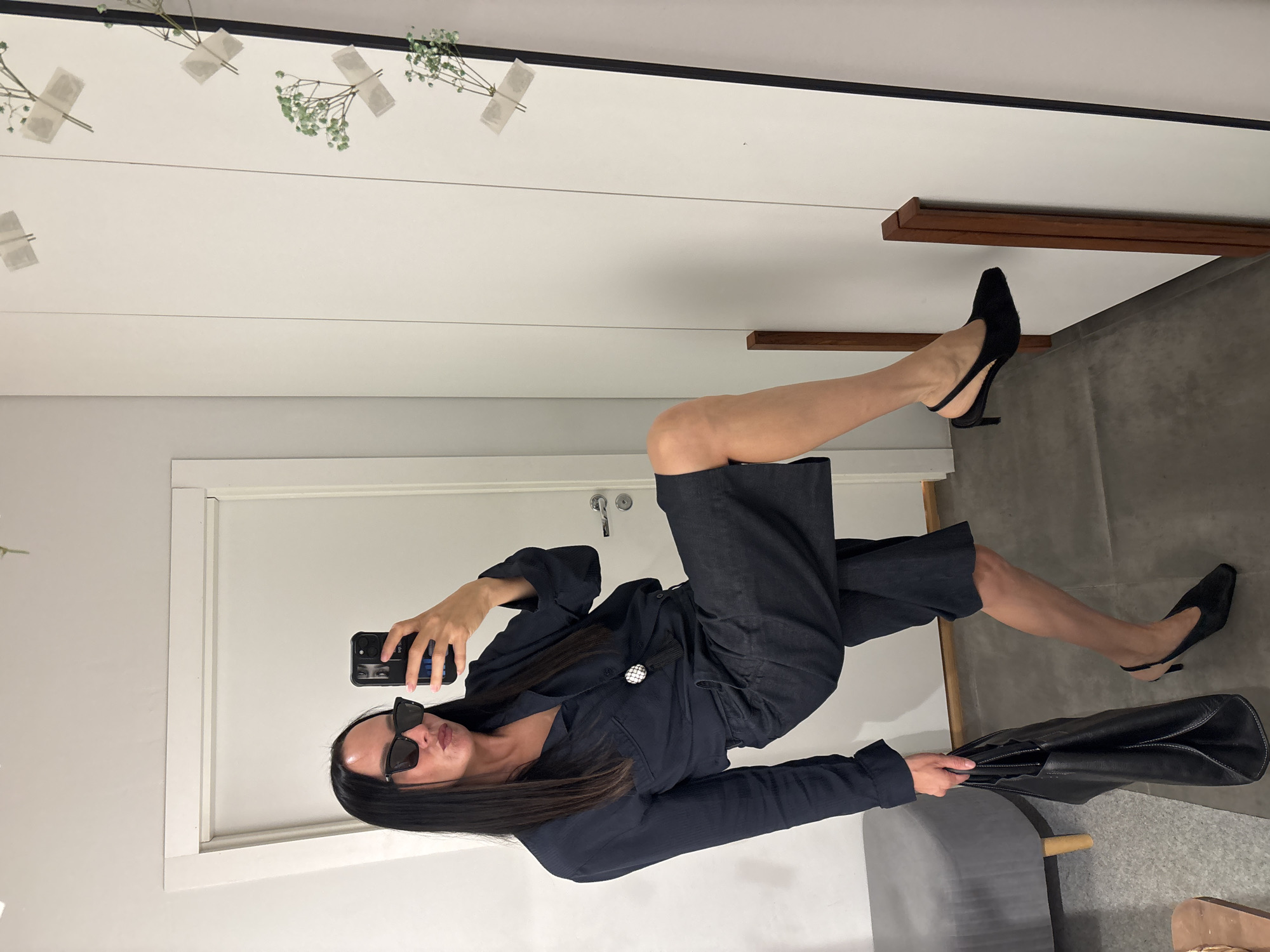 A woman is posing in front of a mirror, taking a selfie with a smartphone. She is wearing a dark blue outfit consisting of a jacket and cropped pants, accessorized with black sunglasses and pointed black heels. She also holds a large black leather handbag. The setting appears to be an interior space, potentially a clothing store dressing room. The background includes a white wall, a door, and some decorations of small white flowers attached to the mirror. I cannot ascertain the precise location in the city of Chaikovsky, Russia, from the visual information provided.