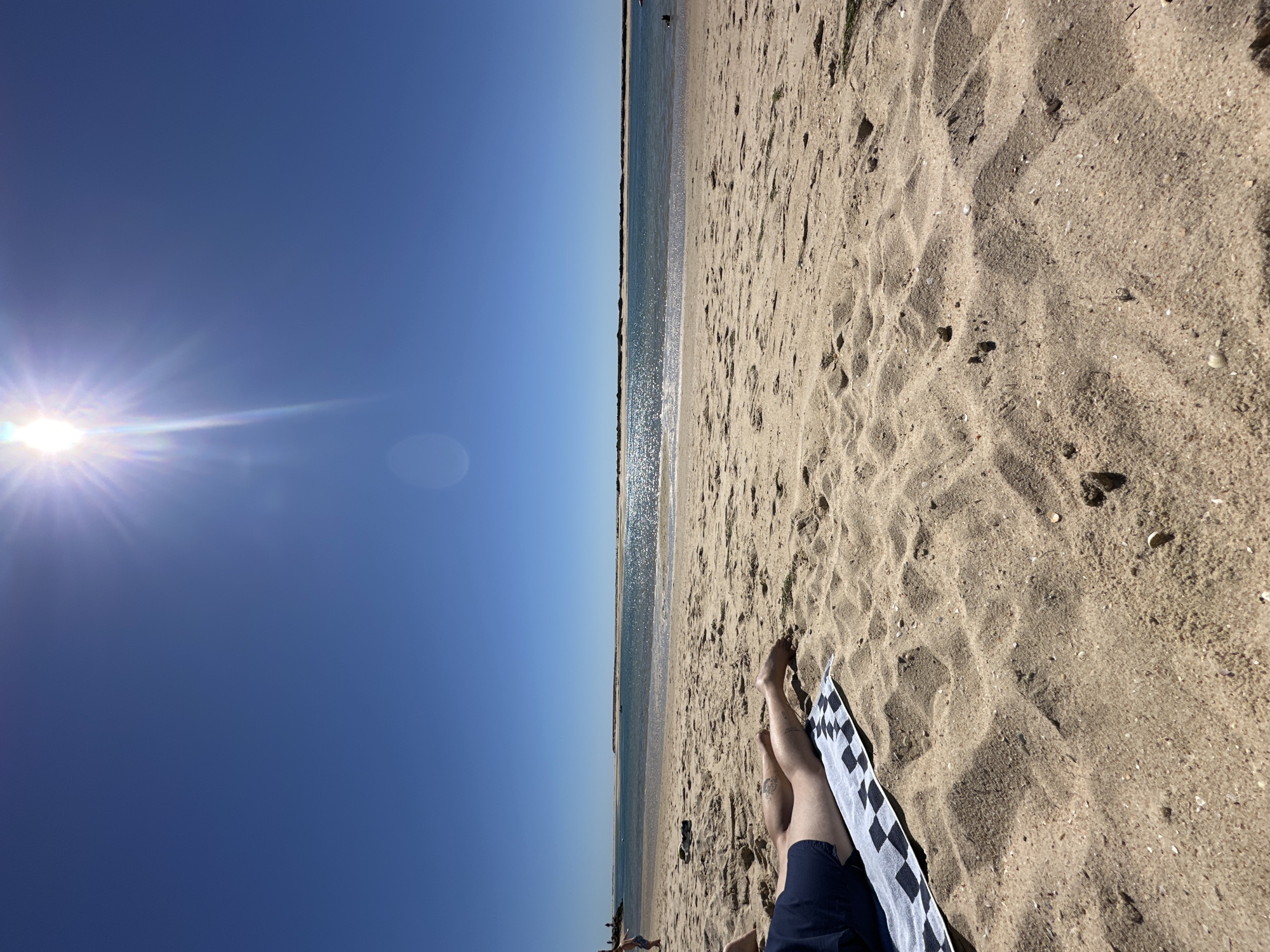 The image presents a beach scene in Olhão, Portugal. A bright sun shines in the upper left corner of a clear blue sky. In the center of the image, a thin strip of water separates the sky from a sandy beach. The beach occupies the right side of the image, exhibiting varying textures and shadows in the sand. In the lower right corner, the legs of a person are visible, resting on a black and white checkered towel. The person is wearing dark blue shorts. In the distance, along the horizon line, there are indistinct figures and structures, likely other beachgoers and possibly buildings.