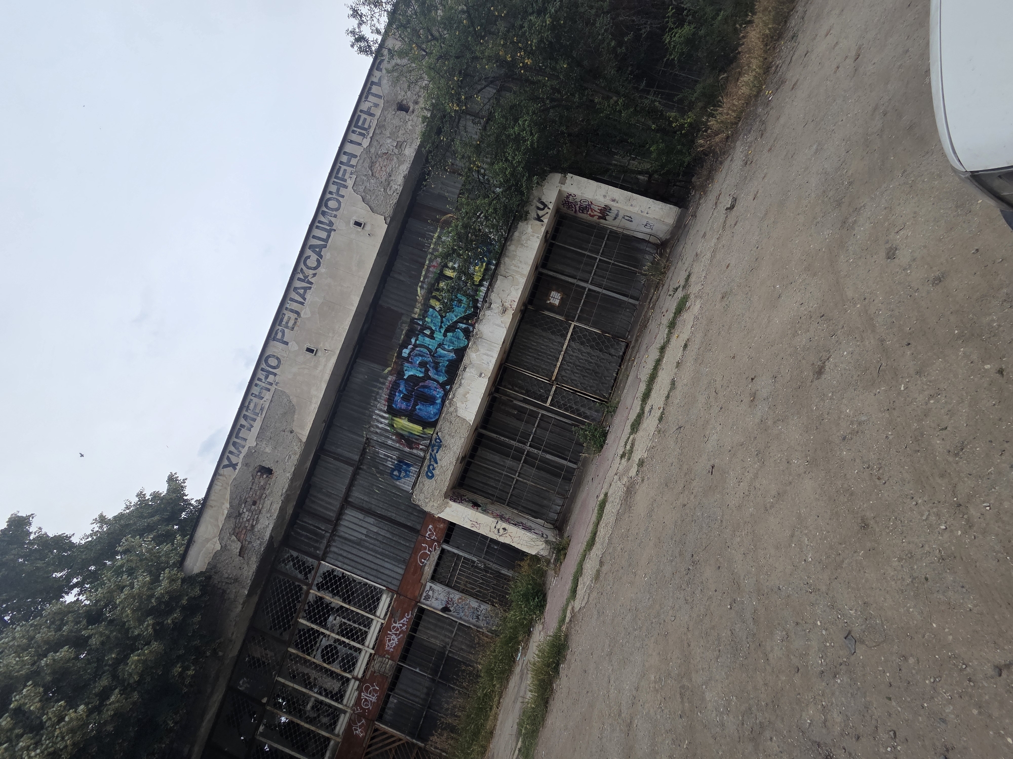 An outdoor, low-angle, wide shot captures a derelict, graffiti-covered building with Cyrillic text on its facade. The building appears to be a former rehabilitation center, as indicated by the partial text "ХИГИЕННО РЕЛАКСАЦИОНЕН ЦЕНТР" (Hygiene Relaxation Center) visible on the upper part of the structure. The building's exterior is constructed from corrugated metal siding and features large, grimy windows with metal security bars. Graffiti art in various colors adorns the lower section of the building, particularly around the entrance. Overgrown vegetation, including trees and tall grass, encroaches on the building, further emphasizing its abandoned state. The ground in the foreground is a dirt and gravel surface, with a hint of a white car roof visible at the bottom right edge of the frame. The sky is overcast and a bird is in flight. The image has a slightly tilted perspective.