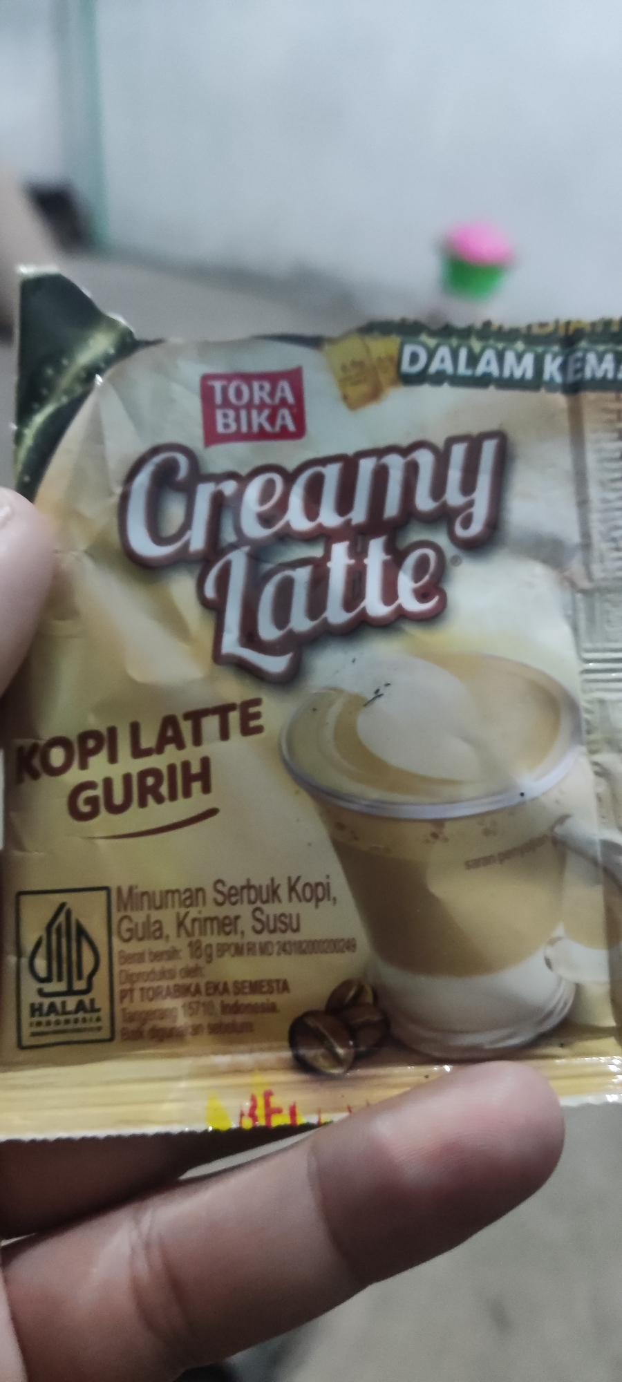A hand is holding a packet of Tora Bika Creamy Latte coffee. The packet is mostly yellow and brown and has text and images on it. The text includes "TORA BIKA", "Creamy Latte", "KOPI LATTE GURIH", and information about the ingredients and manufacturer. The packet also features a Halal certification logo and an image of a coffee cup. The background is blurred and appears to be an indoor setting with some light sources. The image does not provide any information about the city of Karangwangun, Indonesia, or any specific location within it.
