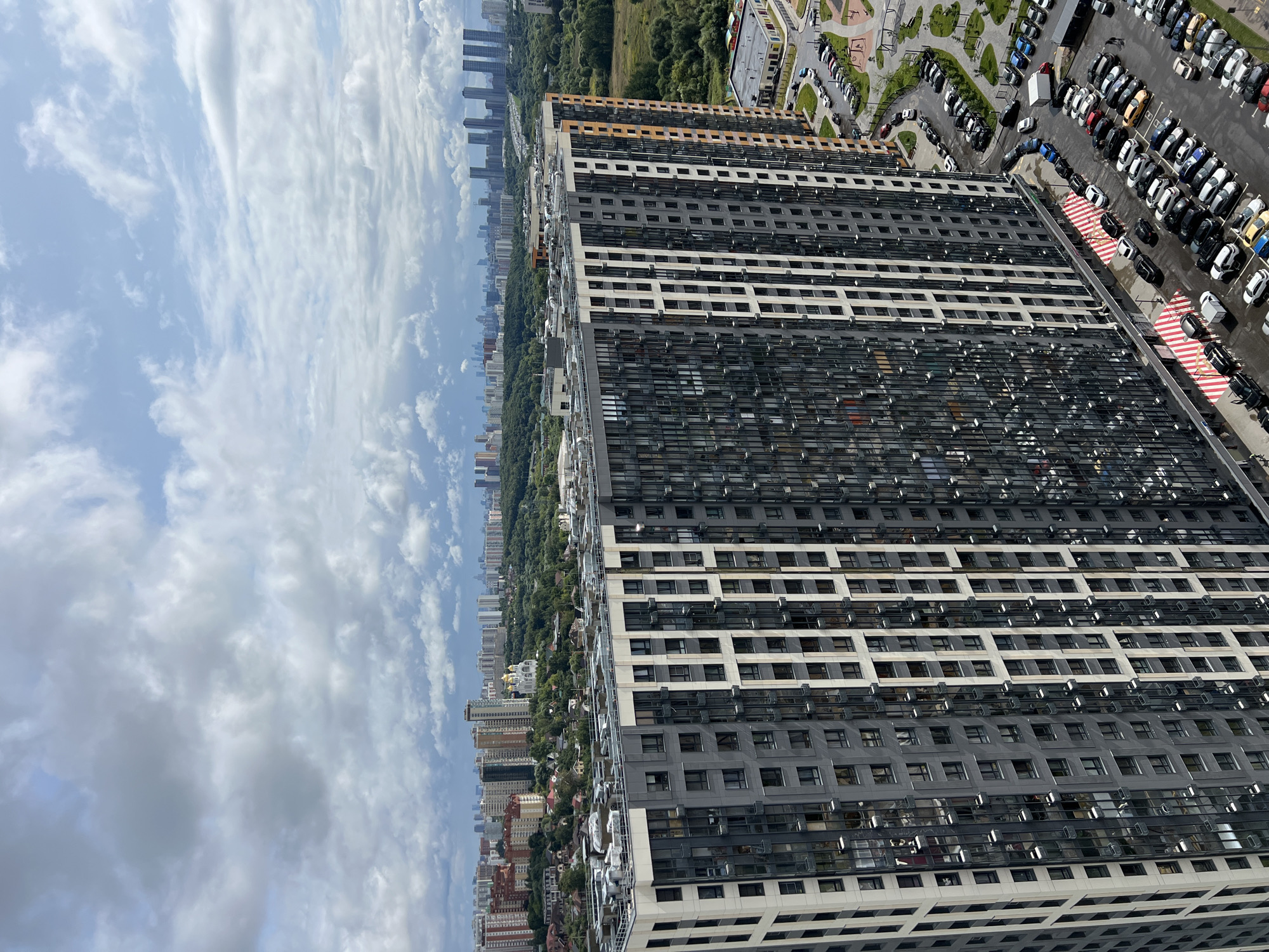 This aerial view captures a modern residential complex in Khimki, Russia. The dominant feature is a tall, multi-story building with a dark gray and white facade, characterized by numerous windows. To the right of this building, a parking lot is filled with many cars. Further in the background, a cityscape unfolds with more residential buildings, a prominent white church with golden domes, and a dense line of trees separating the urban areas from further development. The sky is partly cloudy with a mix of gray and white clouds.