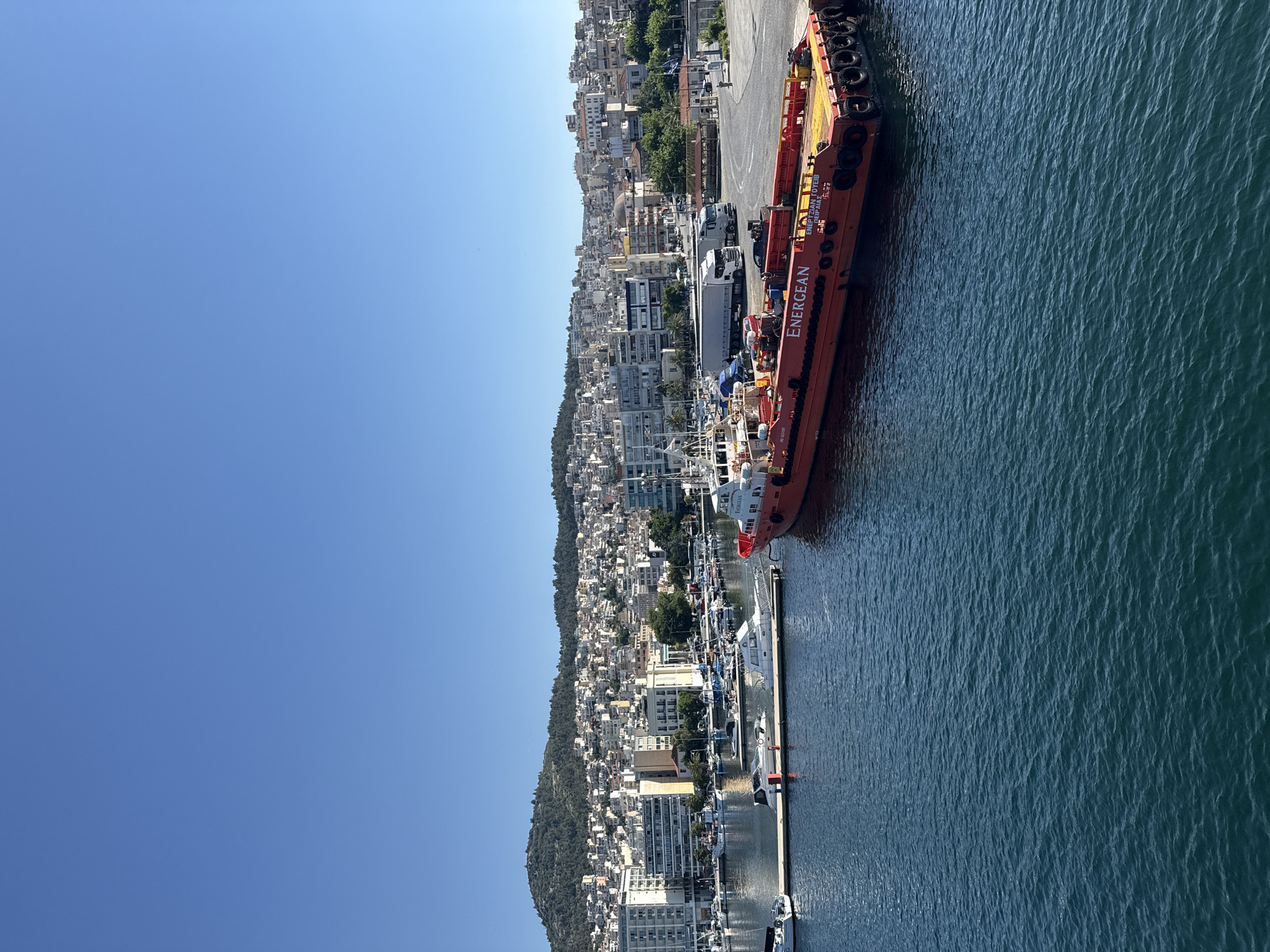 This is a wide shot of a harbor in Kavala, Greece, on a clear, sunny day. The sky is a vibrant blue, with no clouds visible. The water in the foreground is a deep, dark blue with small ripples.
In the harbor, several boats are docked. Closest to the viewer is a large, bright orange barge with "ENERGEAN" written in white lettering on its side. It has yellow accents and a deck covered in equipment. To the left of the barge, a white pier extends into the water, with several smaller white boats docked along it. Further back in the harbor, more boats and yachts of various sizes are visible.
Beyond the harbor, the city of Kavala stretches up a hillside. Buildings of white, beige, and grey are densely packed together, with terracotta roofs visible on some. Green trees are interspersed among the buildings, and the top of a mosque with a dome can be seen in the middle ground. A hilly, tree-covered landscape forms the backdrop to the city, reaching up to the clear blue sky.
The scene appears to be a busy port and a coastal city on a typical summer day.