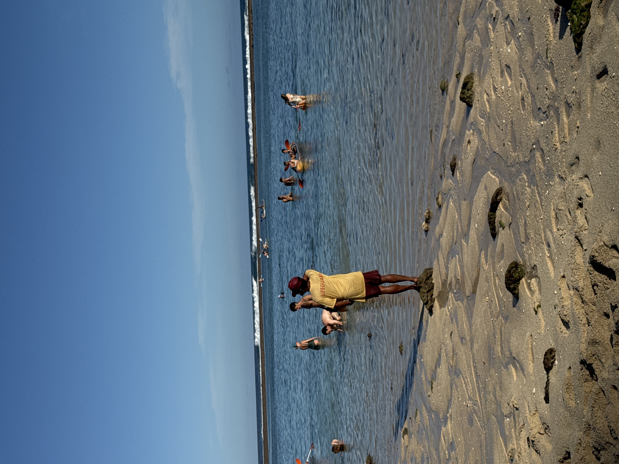 A clear, sunny day at low tide in Kuta Selatan, Indonesia. A lifeguard wearing a yellow shirt and red cap stands on the wet, sandy beach, facing a group of people in the shallow water. Some people are kayaking. Footprints and patterns are visible in the sand. The ocean extends to the horizon, with a faint line of waves breaking in the distance. The sky is a clear blue with a thin strip of white at the edge.