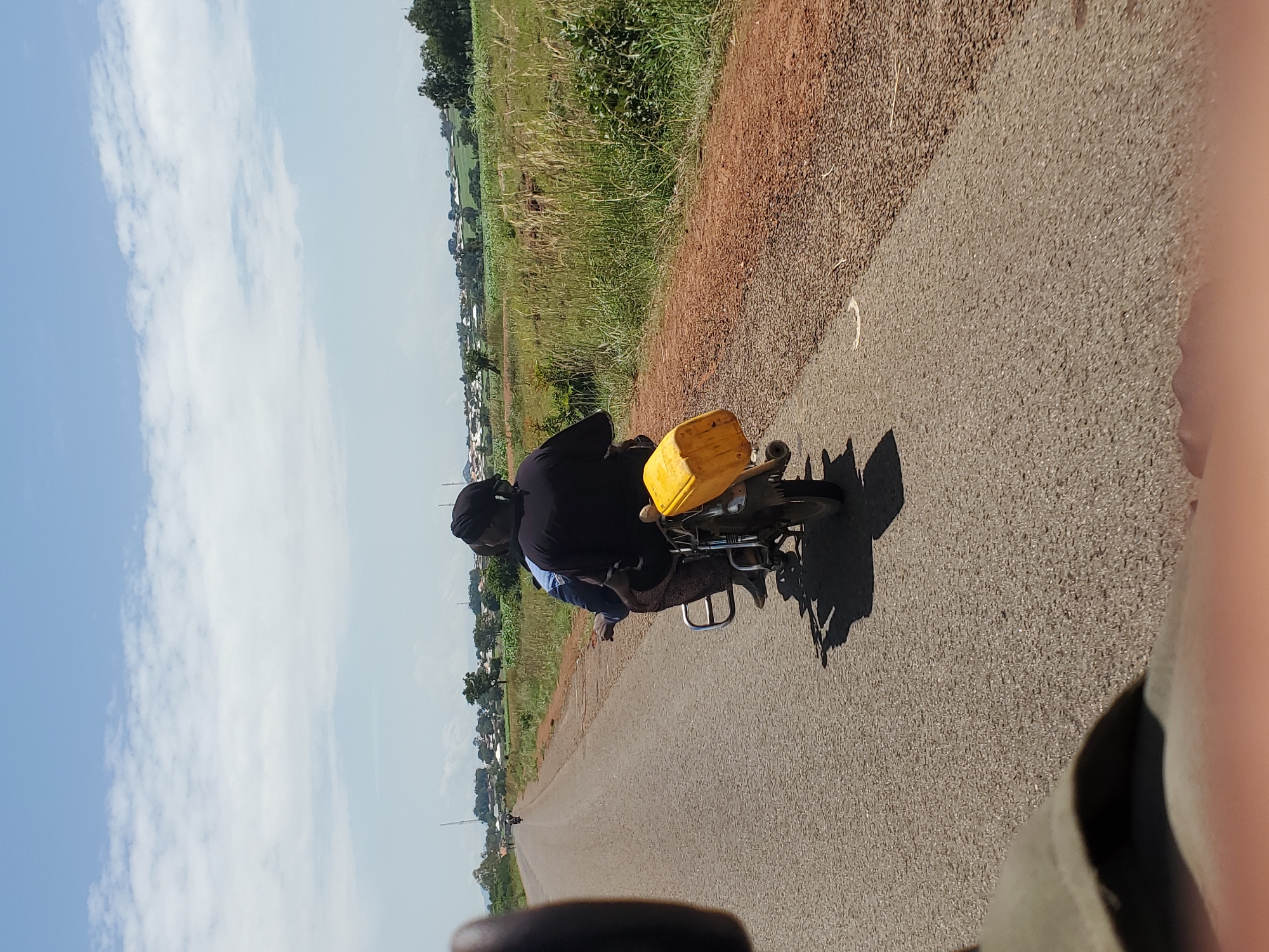 The image depicts a rear view of two individuals on a motorcycle traveling on a paved road. The person in the back is wearing a black shirt and a headwrap, and a yellow container is attached to the back of the motorcycle. The motorcycle is moving away from the camera. The road is flanked by a dirt shoulder on the right, with sparse vegetation. In the background, there is a landscape with fields, trees, and buildings, suggesting a rural or semi-urban environment. The sky is blue with scattered white clouds. The overall scene suggests a sunny day. The image does not contain any visual information that can confirm or deny the location in the city of Pankshin, Nigeria.