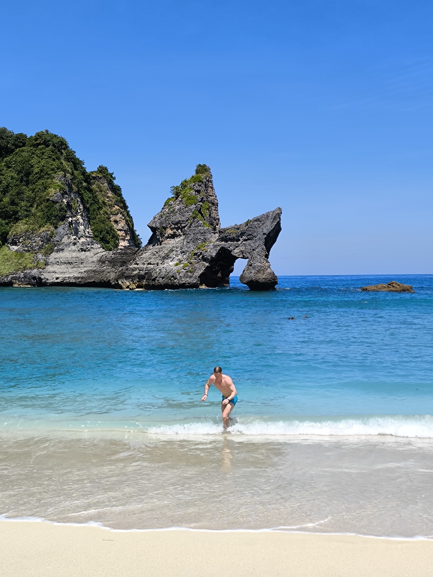 A full shot shows a fair-skinned man in blue swim trunks walking out of the ocean onto the sand at Atuh Beach on Nusa Penida Island in Indonesia on a bright, sunny day. The man has blond hair and is in the middle of the water, walking toward the viewer. The ocean water is a pale blue-green color and is slightly foamy. Behind the man, the ocean is a darker shade of turquoise blue. In the distance, two people are swimming in the ocean. To the left of the man and the swimmers, there is a large landmass of black rock, covered in green vegetation. The rock formation has a large hole in the middle, creating a natural arch. To the right of the rocky landmass is another small island made of the same black rock. The sand is a light tan color and is wet from the ocean water. The sky above is a clear blue color.