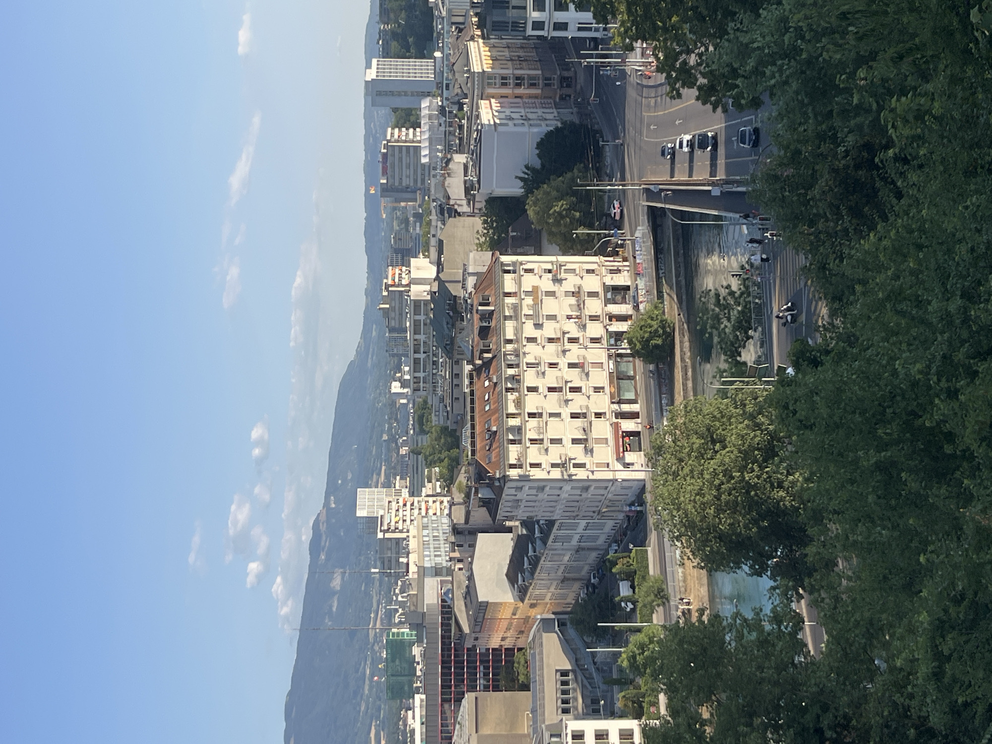This is a vertical, eye-level, medium shot of a city scene in Geneva, Switzerland, taken on a sunny day with a clear blue sky and scattered white clouds. In the foreground, lush green trees partially obscure the view. In the mid-ground, a city scape unfolds with several buildings of varying heights and architectural styles. A notable building in the center is a multi-story structure with a light-colored facade, numerous windows, and balconies, characteristic of European architecture. A river, appearing turquoise in color, flows through the city, crossed by a bridge carrying vehicular and pedestrian traffic. Cars and motorcycles are visible on the road. In the background, more buildings, including modern high-rises, stretch towards a distant mountain range. The overall impression is that of a vibrant, urban environment with natural elements.