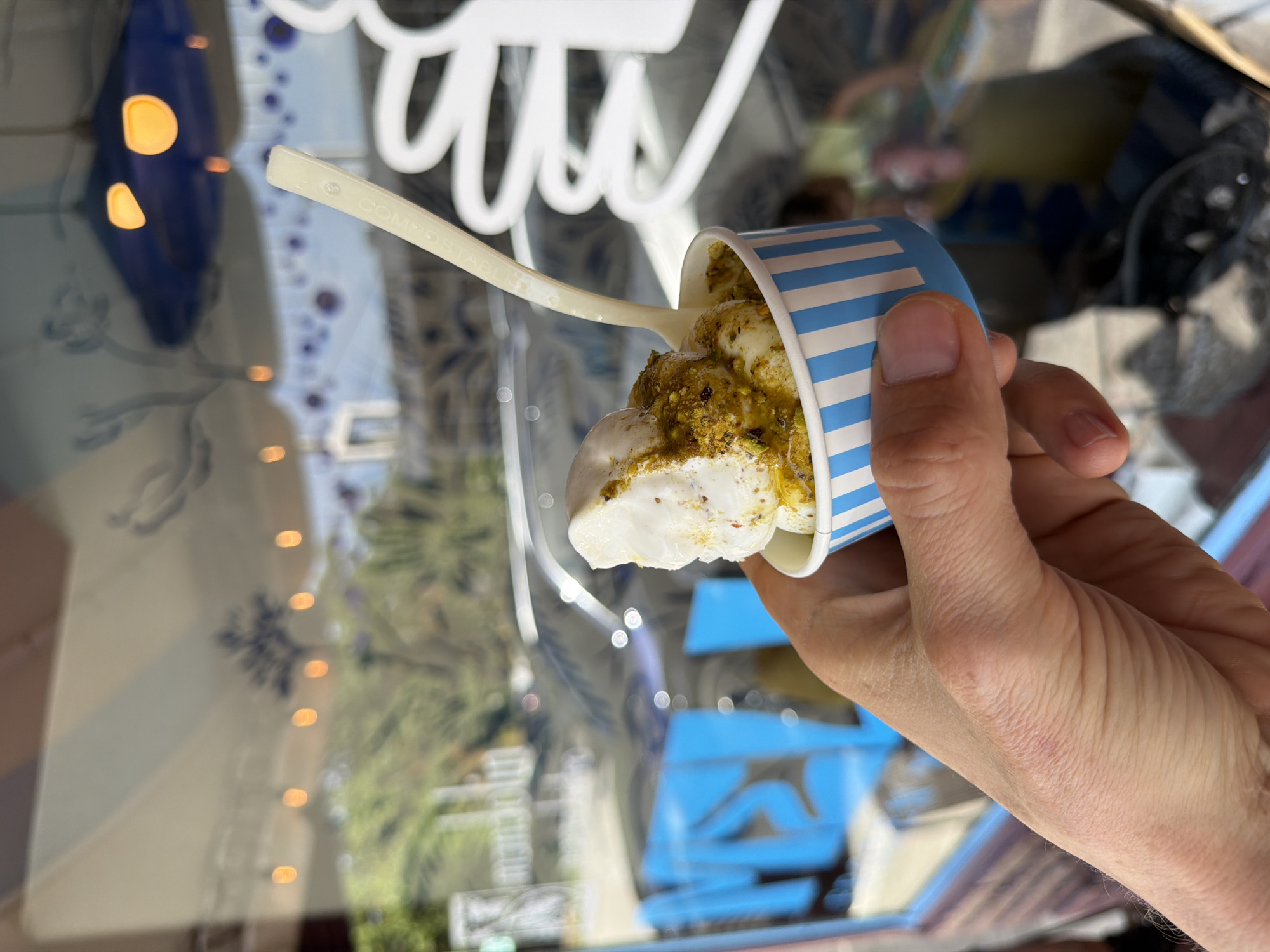 A hand holds a blue and white striped paper cup filled with soft-serve ice cream topped with chopped pistachios. A white plastic spoon is inserted into the ice cream. The scene appears to be outdoors, with a blurred background showing trees, a car, and a building with reflective windows. The overall ambiance suggests a sunny day, possibly in a tourist area. The setting is the city of Miami, United States of America.