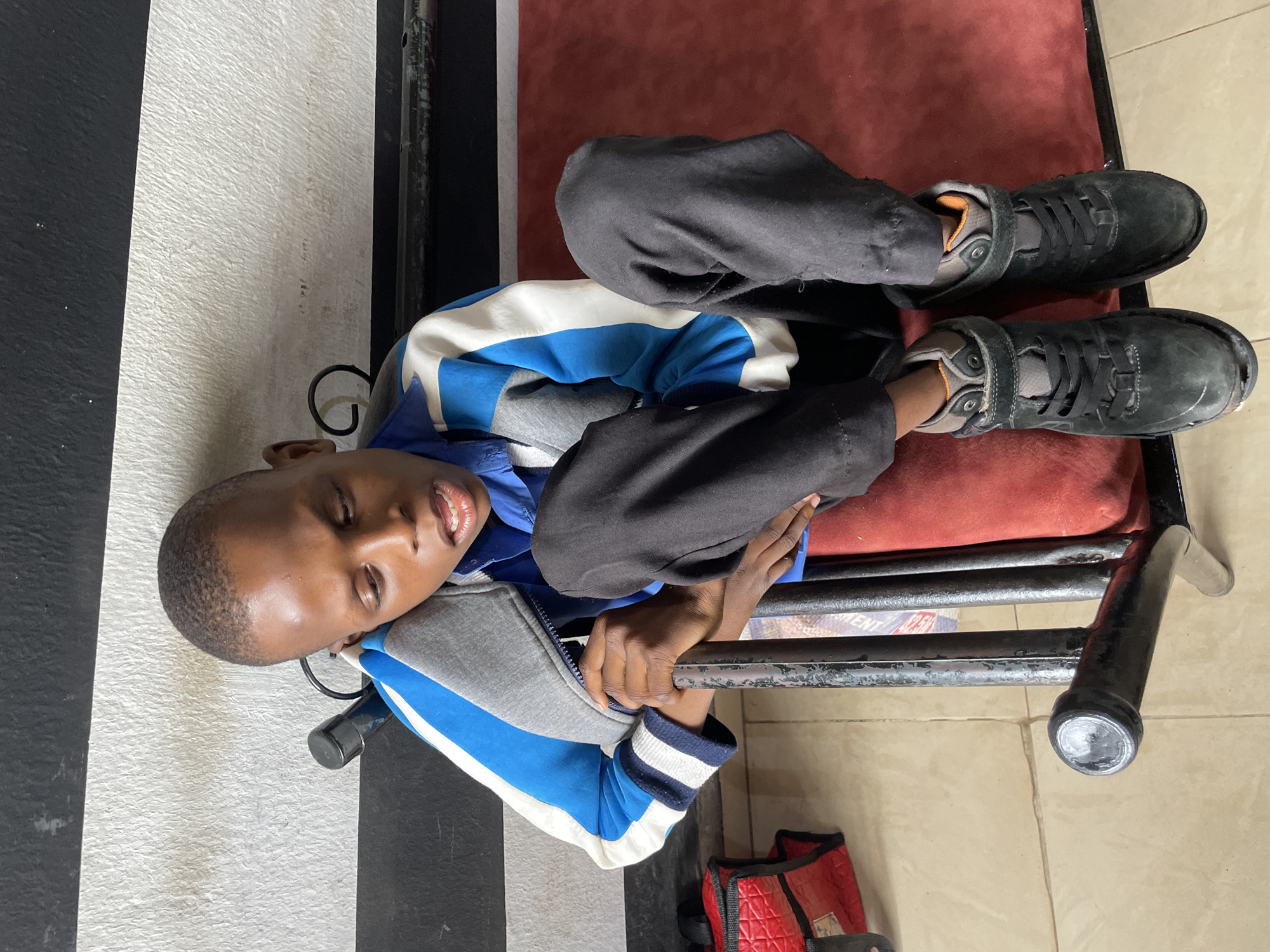 A young boy is lying on his back on a red padded surface, possibly a seat or cushion. He is wearing a blue and white striped jacket over a blue shirt, and black pants. His feet, clad in black lace-up boots with velcro straps, are visible. The boy's eyes are closed, and his mouth is slightly open, giving him a relaxed or possibly sleeping expression.
The background shows a striped wall, with a black and white pattern, and a tiled floor. The setting appears to be indoors, possibly a waiting area or a room in a house. The lighting suggests it is daytime, but the exact time of day is not discernible. There are no obvious weather cues.
A black metal frame is visible, supporting the red padded surface. Part of a red bag with a quilted pattern is on the floor near the boy's feet. Some text is visible on a rolled-up item beneath the red cushion, but it is too blurry to read. The overall impression is of a casual and relaxed moment for the boy. The location context is Jos, Nigeria.