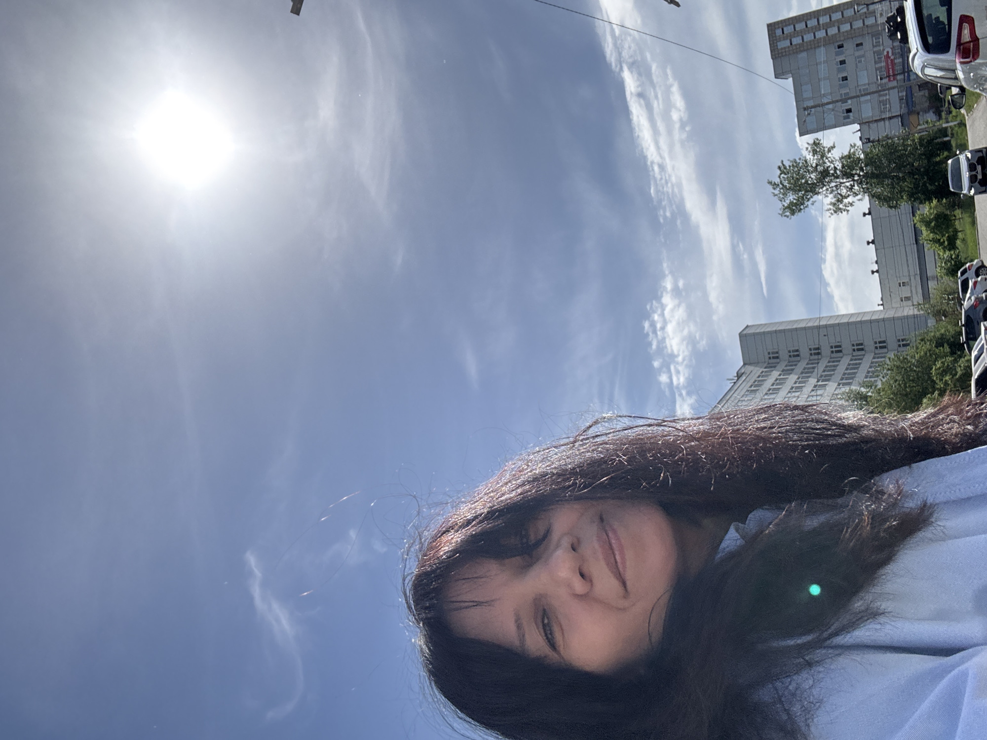 A low-angle, medium shot depicts a woman with dark, shoulder-length hair against a backdrop of a bright blue sky and city buildings. Her hair appears to be blowing slightly in the wind. The sun is visible in the upper left corner of the frame. In the upper right corner, tall buildings are partially visible, along with some trees and parked cars, suggesting an urban environment. The woman is smiling slightly, and the photo appears to have been taken outdoors during the daytime.