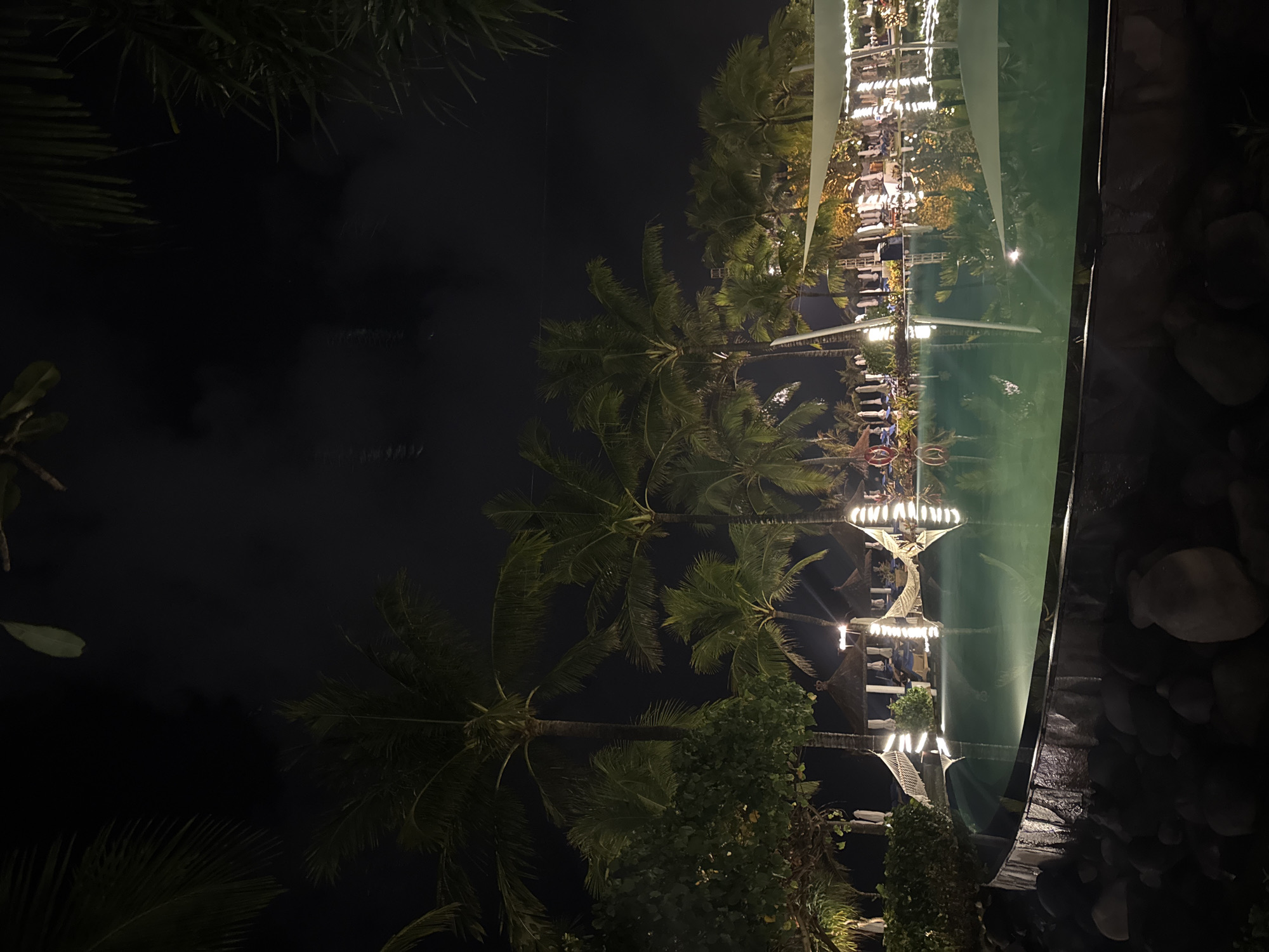 The image depicts a resort swimming pool at night in Kuta Selatan, Indonesia. Palm trees are visible in the background, silhouetted against a dark sky. The pool itself is illuminated by underwater lights, giving the water a green glow. String lights are draped along the edge of the pool and on the surrounding structures. There are several thatched-roof cabanas and lounge chairs visible, with some people appearing to be seated. Hammocks are also visible, suspended between palm trees. The foreground is filled with large, dark rocks. The overall atmosphere is tropical and serene.