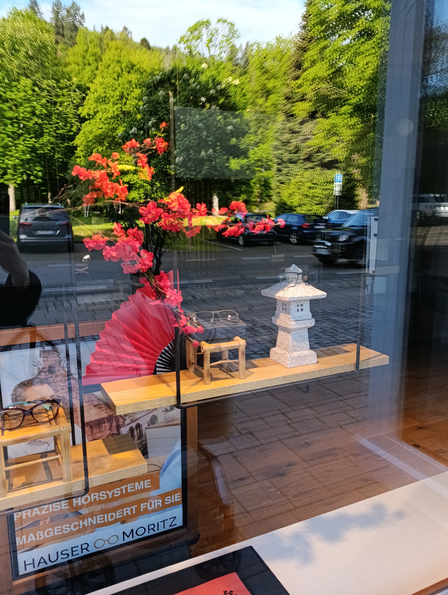 A retail window display of eyeglasses on shelves, including eyeglasses perched atop a small wooden stand and a decorative stone pagoda. The retail location is branded with "HAUSER MORITZ PRAZISE HORSYSTEME MABGESCHNEIDERT FUR SIE", with eyeglasses replacing one of the "o"s in Moritz. Outside the window, a street scene is visible with parked vehicles and lush green trees.