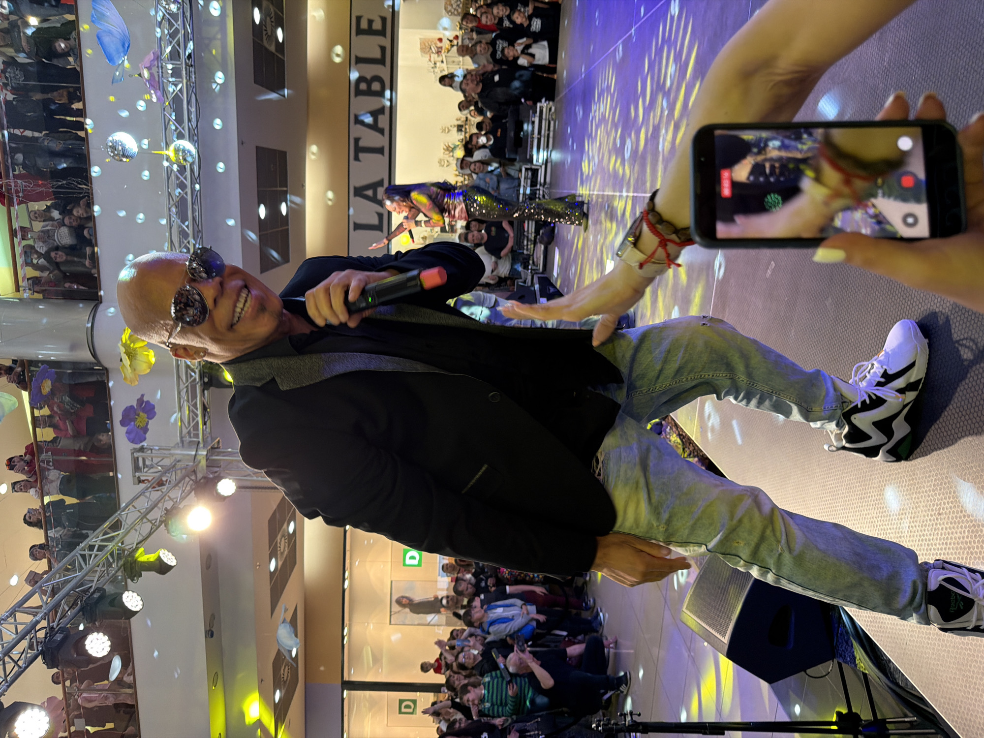 On a stage inside what appears to be a shopping mall in Pleven, Bulgaria, a bald, middle-aged man in a black blazer, light-wash jeans, and black-and-white athletic shoes smiles broadly while holding a microphone. He is wearing sunglasses and is slightly leaning back. Another person on stage, visible behind him, is wearing a shimmering outfit. In the foreground, someone holds up a smartphone to record the performance. Behind the stage, an audience watches. A store sign reading "LA TABLE" is visible in the background. The stage is lit with yellow and purple lights, and decorations including mirror balls and artificial flowers hang from the ceiling. Another part of the crowd is visible on a raised section, and some people are standing along a glass barrier on another level of the mall. The floor of the stage has a hexagonal pattern. A speaker sits near the front of the stage, close to the person holding the phone.