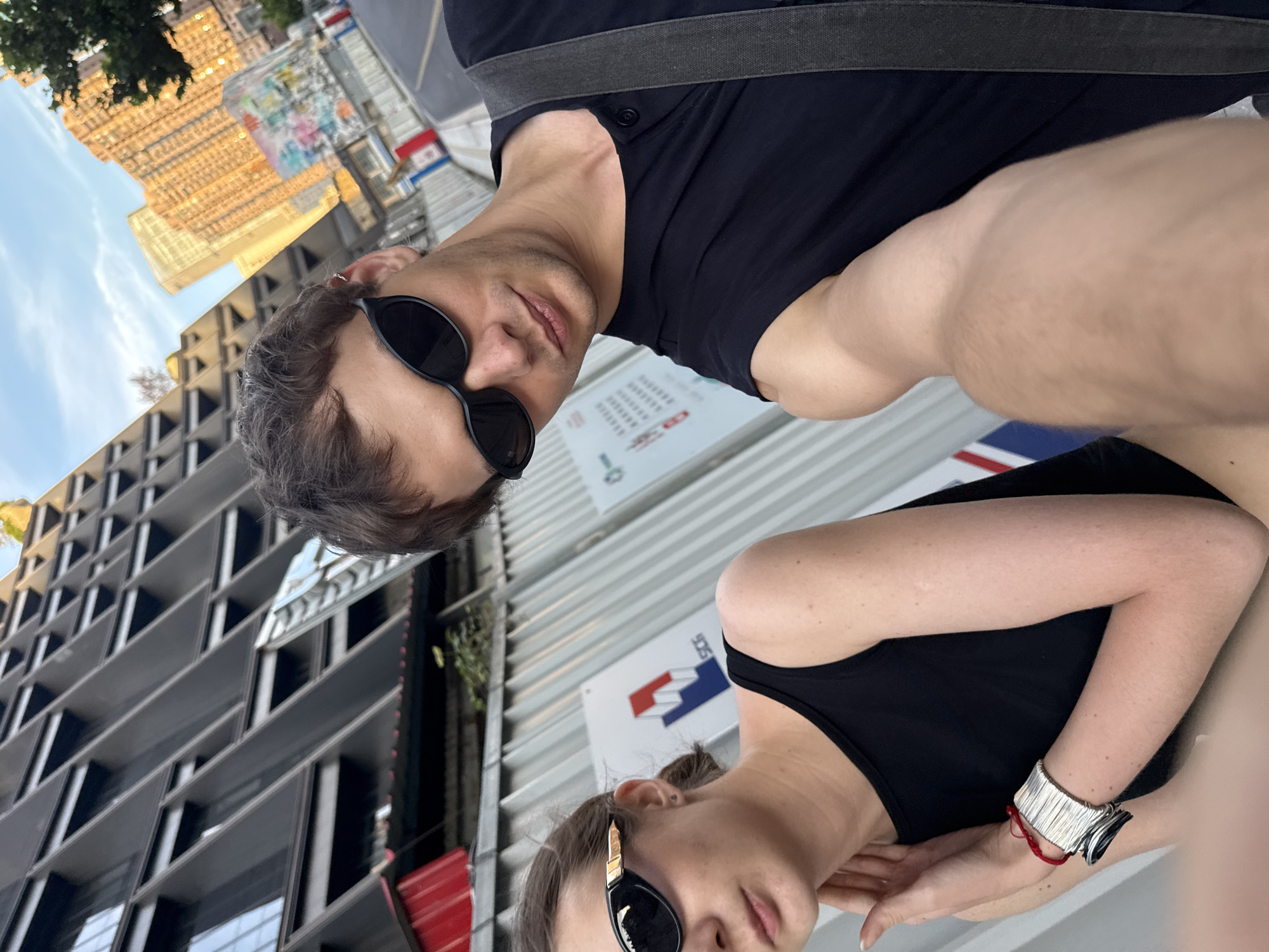 This image is a dynamic selfie featuring two individuals, taken at an acute angle that rotates the scene 90 degrees clockwise. The setting is an urban environment, identified as Putuo District, China, characterized by modern architecture and ongoing construction.
In the foreground, a male with dark hair and stubble wears black sunglasses and a black t-shirt with a dark strap across his shoulder, possibly from a bag. He looks directly towards the camera with a neutral expression. Behind him and slightly below, a female with light brown hair, pulled back, also wears black sunglasses and a black tank top. Her right arm is visible, adorned with a distinctive wide, textured silver cuff bracelet and a red string bracelet underneath. She looks slightly past the camera.
The background reveals a bustling city scene under a bright sky with sparse clouds, indicating daytime. Tall buildings with contemporary designs dominate the view. One prominent building exhibits a grid-like facade with numerous small, possibly reflective, square or rectangular windows/panels that catch the sunlight. Another building has a darker, more angular and geometric design. Construction barriers or temporary fences run across the middle ground, separating the subjects from the distant structures.
Several signs are affixed to these barriers. One sign prominently displays a logo featuring a stylized blue mountain peak over a red base, resembling an "M". Other signs have text, though it is largely illegible due to the distance and angle, with some appearing as rows of asterisks or numerical sequences. The overall impression is one of candid urban exploration or a casual outing in a modern city setting.