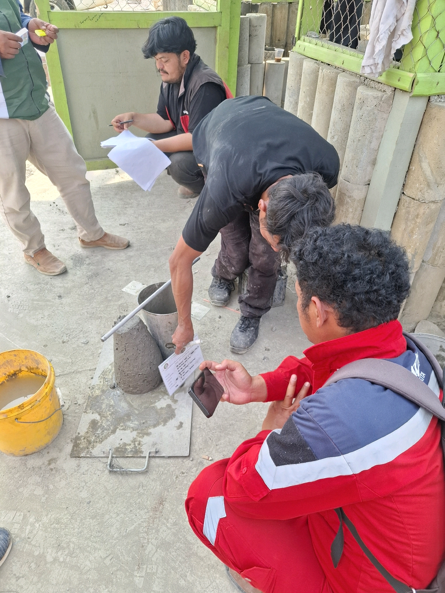 In Cijangkar, Indonesia, three individuals are engaged in a concrete slump test. One person, wearing a green vest and beige pants, stands holding a yellow marker. A second person, crouched and wearing a black vest over a black shirt and dark pants, holds a pen and paper. The third individual, kneeling and wearing a red and blue uniform, is holding a phone and pointing at a concrete cone. The environment consists of a concrete ground, a yellow bucket, a metal bucket, concrete cylinders, and a green metal fence. The scene suggests a construction or engineering context where concrete quality is being assessed.
